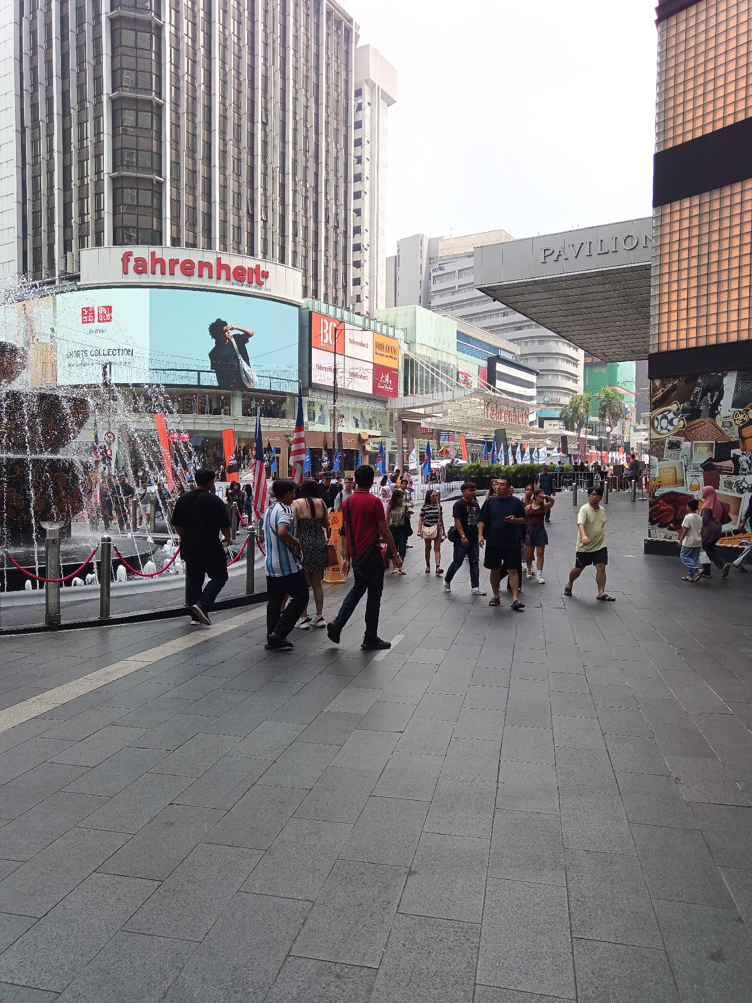 The image depicts a bustling street scene in front of the Pavilion Kuala Lumpur, a prominent shopping mall located on Bukit Bintang, Kuala Lumpur, Malaysia. The foreground is dominated by a wide, gray paved pedestrian walkway. A large, multi-tiered fountain with cascading water is to the left, bordered by a red rope. Behind the fountain, a large digital billboard displays advertisements, including the "fahrenheit" logo. Several people are walking in various directions across the walkway.
In the middle ground, numerous individuals, a mix of locals and tourists, are seen strolling, some in groups, others individually. They are dressed in casual attire, appropriate for a warm climate. Flags, including Malaysian flags, are visible, suggesting a possible event or celebration. The background features the impressive architecture of the Pavilion Kuala Lumpur, with its distinctive glass facade and the prominent "PAVILION" signage. Adjacent buildings and structures of varying heights, typical of a dense urban environment, are also present. The sky is overcast, indicating a cloudy day. The overall atmosphere is one of lively urban activity, with shopping, leisure, and pedestrian movement at the forefront.