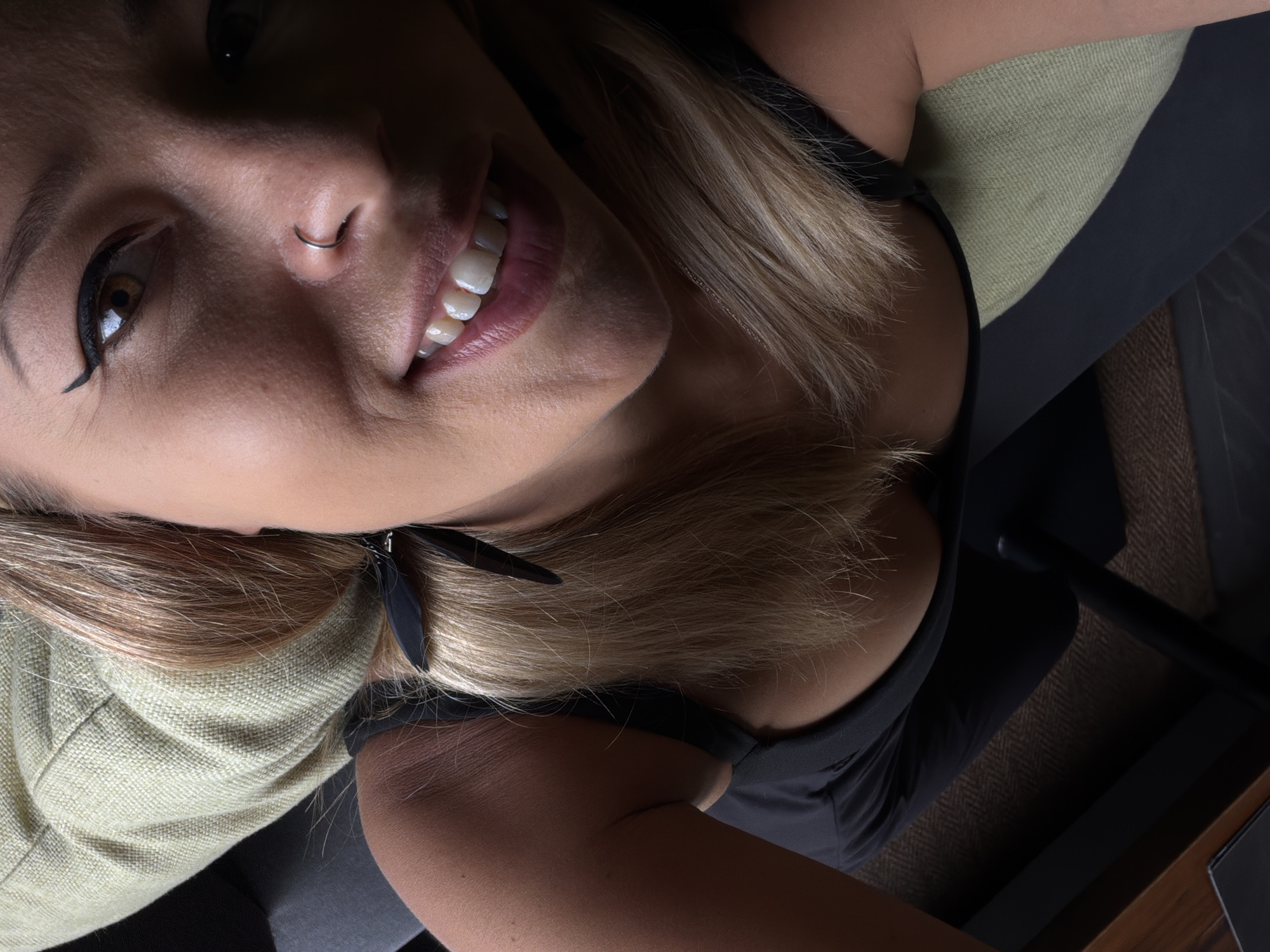 The image is a close-up, high-angle shot of a woman's face and upper body. She has blonde hair, is wearing a dark top, and has makeup on, including winged eyeliner and lipstick. She is also wearing a nose ring. She appears to be indoors, possibly on a couch or chair, with some textured fabric visible in the background. There is no indication of the location being in ตำบลเกาะพะงัน, Thailand.