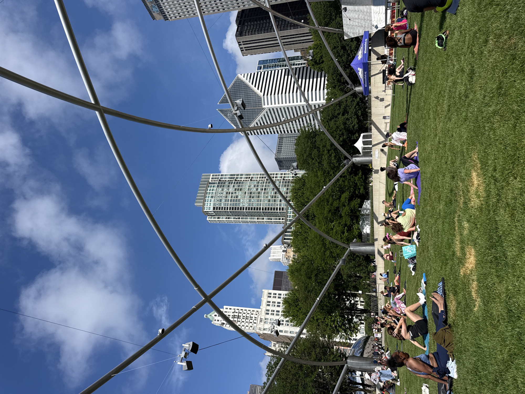 The image depicts a sunny day in Millennium Park, Chicago, Illinois, United States of America, with a clear blue sky dotted with fluffy white clouds. A large, modern metal trellis structure dominates the foreground, arching over a grassy lawn where a group of people are engaged in what appears to be a yoga or exercise class. The individuals are spread out on yoga mats, some in seated or reclining poses, while others are in motion. The backdrop reveals the iconic Chicago skyline, featuring various architectural styles of skyscrapers, including the distinctive geometric white building in the center. Lush green trees are interspersed between the buildings and the park's structures, adding to the urban oasis setting. A blue tent with "MillenniumPark.org" printed on it is visible on the right side of the frame, suggesting an organized event. The overall scene conveys a sense of community activity and urban recreation on a pleasant day.
