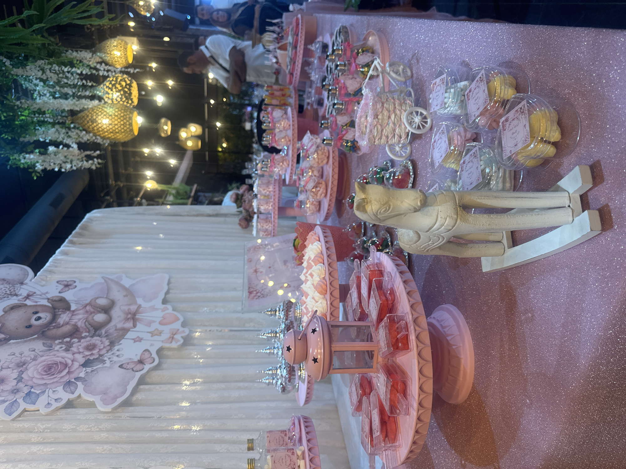 The image depicts a festive dessert table setup, likely for a baby shower or a similar celebration, with a prominent pink and pastel color scheme. The table is covered with a sparkling pink tablecloth.
On the table, various sweets are arranged on tiered pink stands. These include small candies in clear bottles with pink ribbons, what appear to be meringues or macaroons, and individually packaged pink desserts in clear plastic containers. A central element on one of the stands is a pink lantern with star cutouts. A decorative wooden rocking horse is placed in the foreground, serving as a focal point. In the background, a large cutout of a teddy bear sitting on the moon, adorned with roses and butterflies, is visible against a white draped curtain. Hanging from the ceiling are decorative lights and floral arrangements, suggesting an indoor event space.
To the right of the table, several adults are partially visible, engaged in conversation. One man in a white shirt is facing away from the camera, and a woman in a dark top is to his right. The background suggests a dimly lit venue with more ambient lighting and some greenery.
While the provided text mentions "Migdal HaEmek, Israel" in relation to the original image, this specific visual information cannot be directly inferred from the depicted scene itself. The image focuses solely on the decorated dessert table and the immediate surroundings.