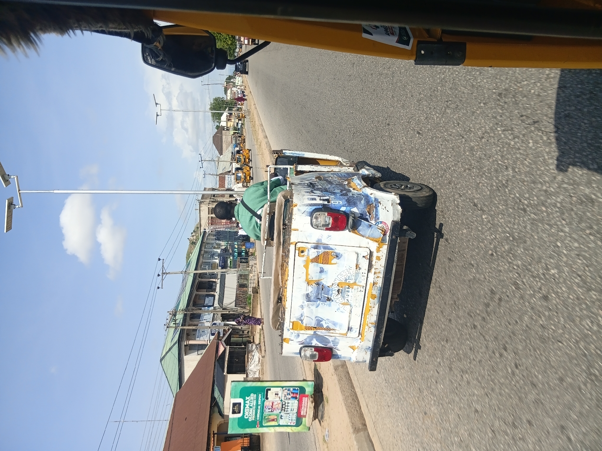 In Jalingo, Nigeria, on a clear day, a person in a green shirt is driving a white, heavily decorated, open-air three-wheeled vehicle down a paved road. The vehicle has a weathered appearance with various decals and paintwork, including images and text that are partially obscured. The driver is seated in the front, and the back of the vehicle is open. The vehicle is moving on the right side of the road, indicated by the direction of traffic and the presence of other vehicles further ahead, including yellow auto-rickshaws.
The environment suggests a commercial or residential area. On the left side of the road, there are buildings with shops, including one with a prominent green sign for "OKEMAX GLOBAL ALLIED PRODUCTS LTD CHEMICAL DEPOT." Further down the road, more buildings with green roofs and balconies line the street. Electrical wires are strung overhead, and a few lampposts are visible. The road itself is asphalt, with painted lane markings and shadows cast by the vehicles and overhead structures. A few pedestrians are visible on the sidewalk in the distance. The scene captures a typical urban street in Nigeria, characterized by its active street life and local transportation.