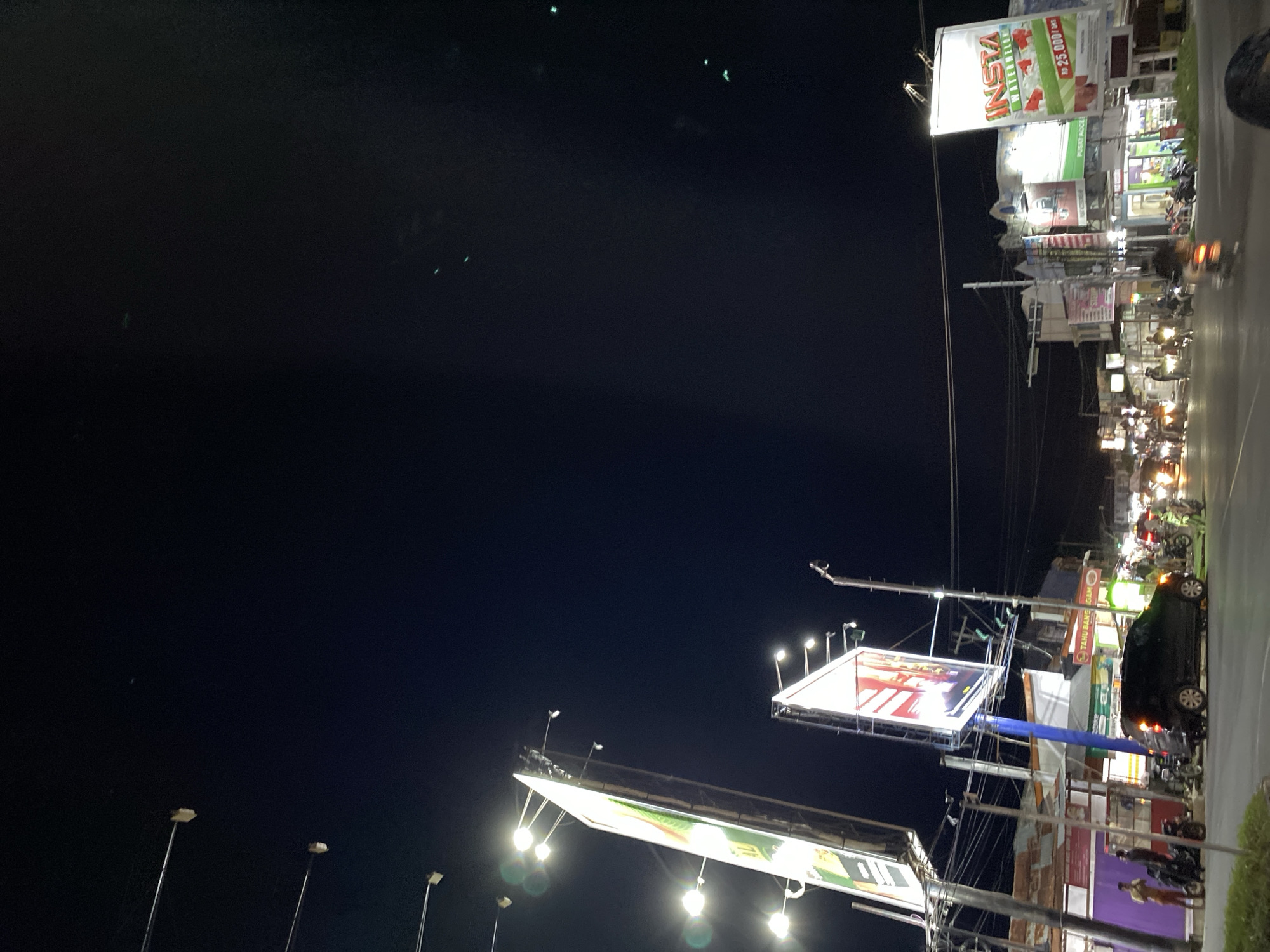 This is a nighttime photograph, likely taken from a moving vehicle, capturing a street scene illuminated by artificial lights. The image is oriented vertically, making it appear as if the viewer is looking upwards from a low angle.
The primary subjects are a series of illuminated billboards and streetlights. One large billboard prominently displays the word "INSTA" in bold red lettering, with "WATERWELL" below it in green. A price of "Rp 25.000 / pcs" is also visible. Other billboards, though less clear, suggest commercial advertisements. Numerous streetlights, some with multiple bulbs, dot the scene, casting pools of light.
In the background, buildings with lit windows and storefronts line the street. Several motorcycles are parked or in motion, indicated by their lights. A dark-colored car is parked on the right side of the road. A few figures, likely pedestrians, are visible near the buildings and motorcycles.
The overall setting appears to be an urban or semi-urban environment at night. The presence of billboards, streetlights, and vehicular traffic points to a commercial or bustling area. The darkness of the sky and the bright artificial lights suggest it is either late evening or night. The reflective wetness on the road surface could indicate recent rain or a damp environment. The image conveys a sense of activity and commerce, even in the darkness.