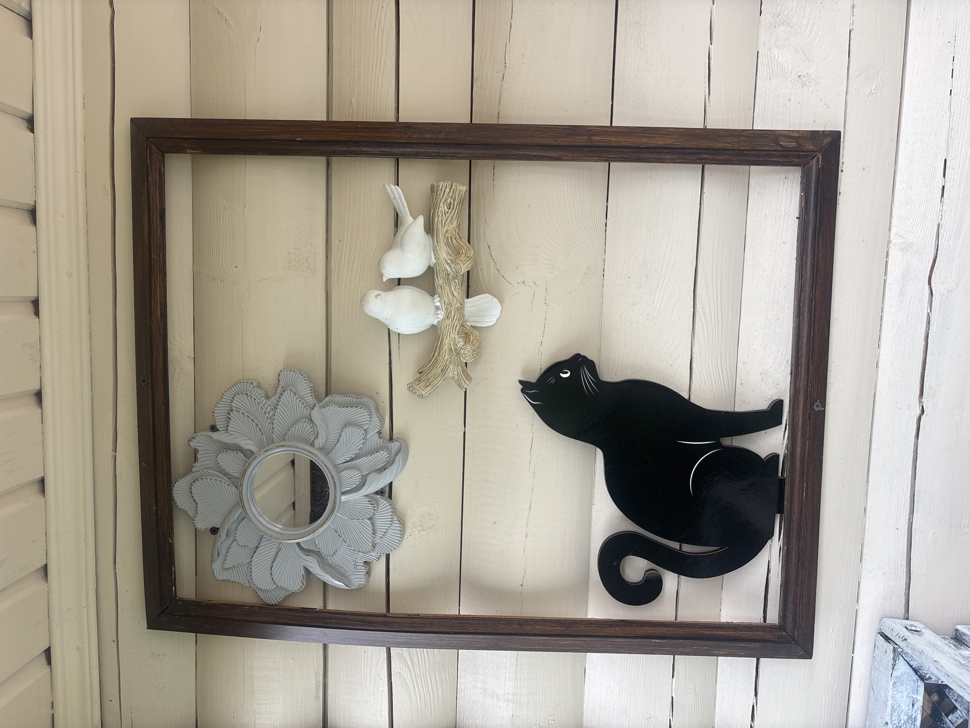 A dark brown wooden frame hangs on a weathered, pale yellow wooden wall, possibly the exterior of a building in Лотос Снт (марьино), Russia. Inside the frame, on the wall, are several decorative items. At the top left, a light gray decorative mirror shaped like a flower is mounted. To the right of the flower mirror, a faux branch with two small white bird figures is attached to the wall. Below the birds, a black silhouette of a cat is affixed to the wall, positioned at the bottom of the frame. The wooden wall exhibits horizontal planks with visible grain and some signs of wear. In the lower left corner, a portion of a white, weathered object, possibly a box or planter, is visible.