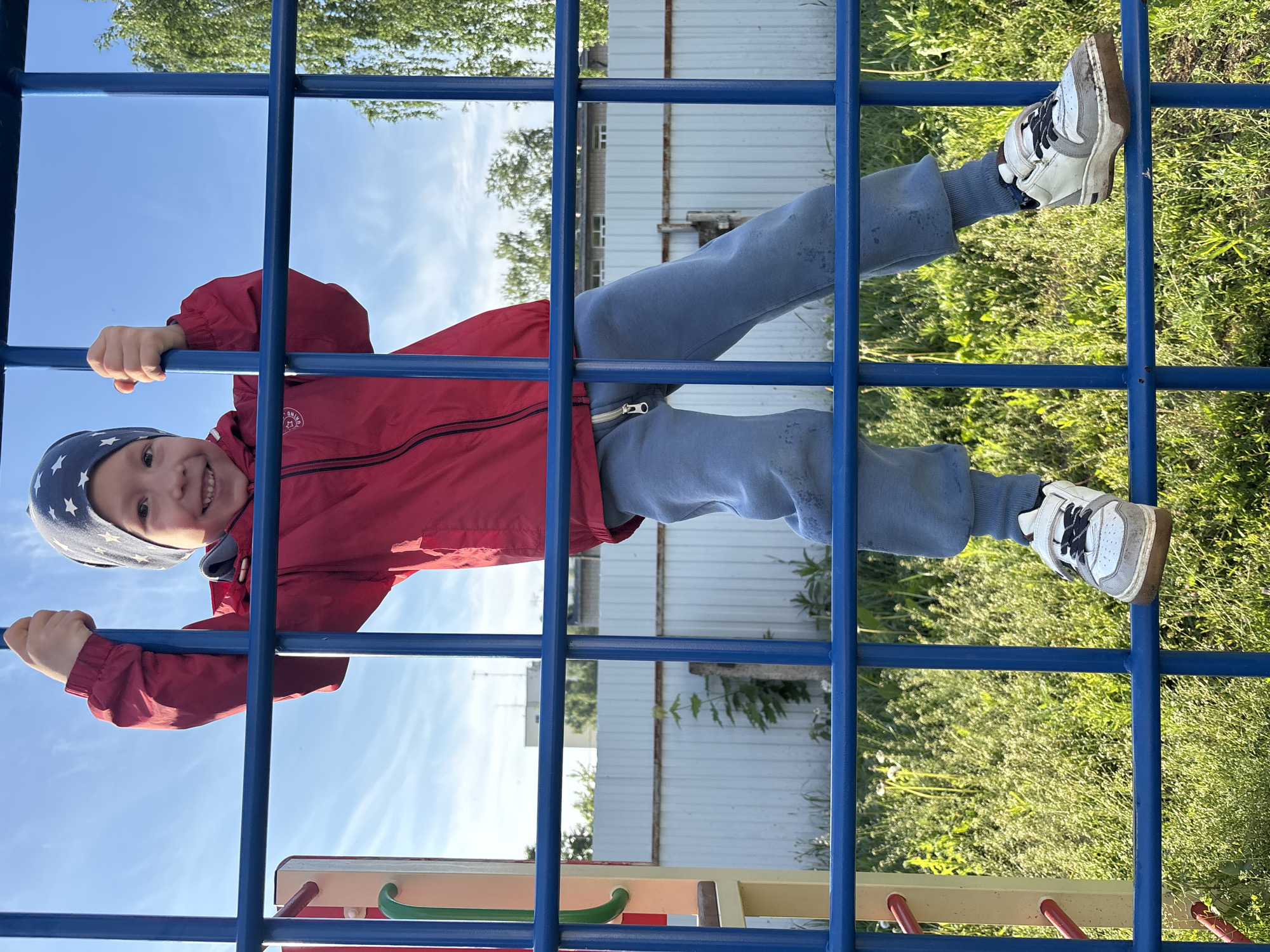 A young child is captured in an outdoor setting, playfully hanging upside down on a blue metal climbing frame, or monkey bars. The child, likely a boy, is wearing a bright red hooded jacket, light blue sweatpants, and a dark blue beanie patterned with white stars. He is smiling broadly at the camera, conveying a sense of joy and excitement.
The perspective is from below, looking up through the bars of the playground structure. The child's hands grip a lower bar, while his feet, clad in white and grey sneakers featuring a visible Nike swoosh, are hooked over an upper bar, securing his inverted position. A small, circular logo, possibly reading "ONIKO," is visible on his red jacket.
The background reveals a clear blue sky with a few wispy clouds, suggesting bright, sunny weather. Lush green grass and various foliage are visible around the play area, along with part of a light-colored building or fence. At the very bottom of the frame, elements of other colorful playground equipment, including yellow, red, and green bars, can be seen. The scene depicts a vibrant and active moment of childhood play.
Location context: Chaikovsky, Russia.