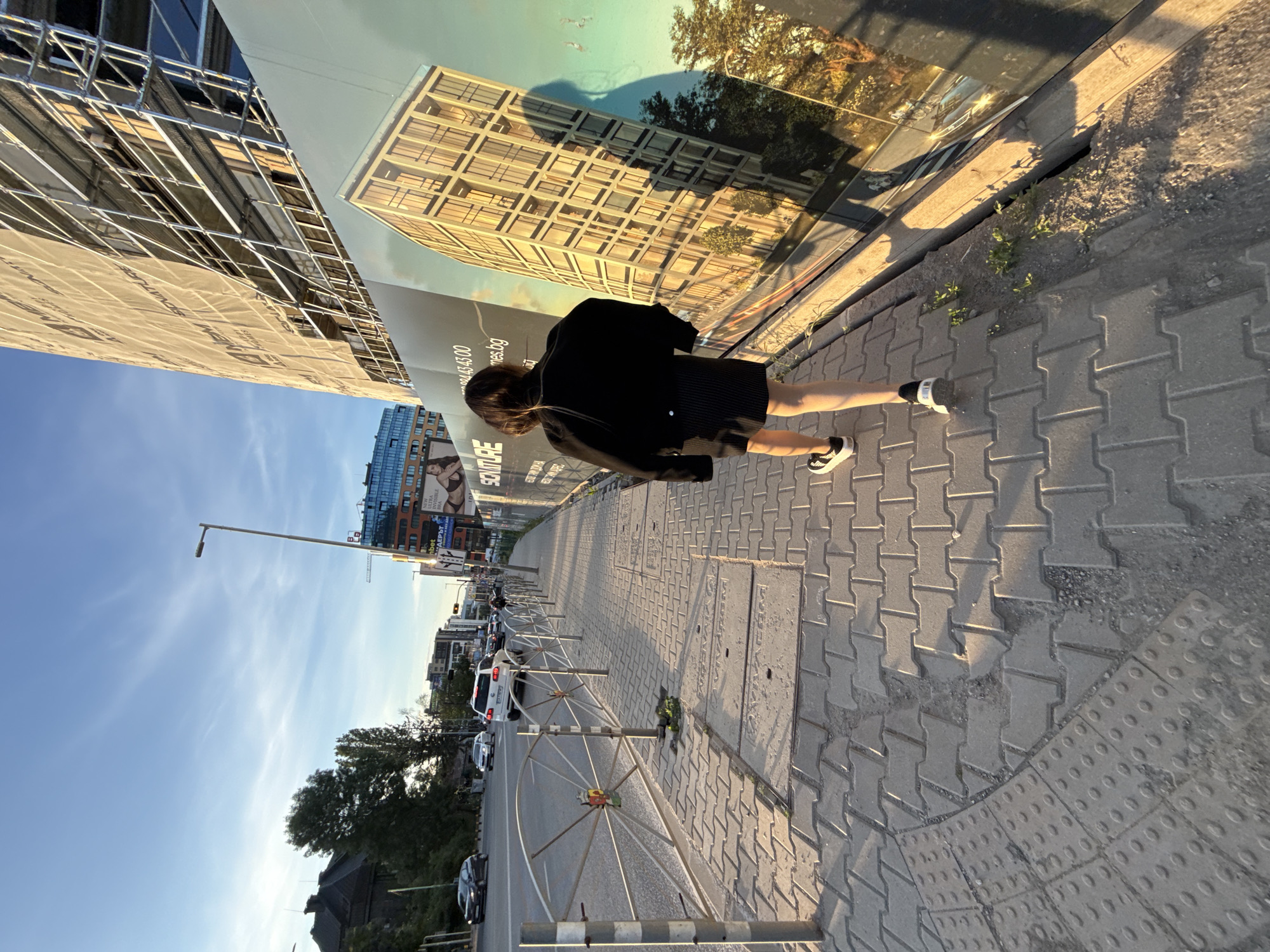 A medium outdoor shot depicts a woman walking on a gray brick sidewalk adjacent to a street in Sofia, Bulgaria. The woman has short brown hair, a black jacket, and a striped skirt and is walking towards the top of the frame away from the camera. The sidewalk extends to the right and curves into a safety barrier of metal rings and poles along the street's edge. A busy street with cars parked and in motion runs parallel to the sidewalk.
In the background, on the left, scaffolding is visible along with a building under construction. A large advertisement featuring a rendered image of a building is mounted on the construction site.
The sky is blue with light clouds. The time of day is likely late afternoon as there are long shadows.
The image appears to have been taken at an angle.