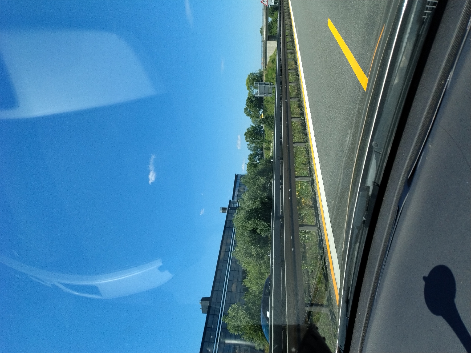 The image captures a view from inside a vehicle, likely on a highway, with a clear blue sky overhead and a few scattered clouds. In the distance, a modern-looking building with extensive glass windows is visible, surrounded by lush green trees. A highway guardrail runs parallel to the road, with a yellow line marking the lane. The scene appears to be set in the city of Hengersberg, Germany, given the context provided. There are no people or explicit actions depicted, but the presence of the guardrail and road markings suggests ongoing traffic. The overall impression is one of a bright, clear day, possibly during a journey through the German countryside or outskirts of a city.