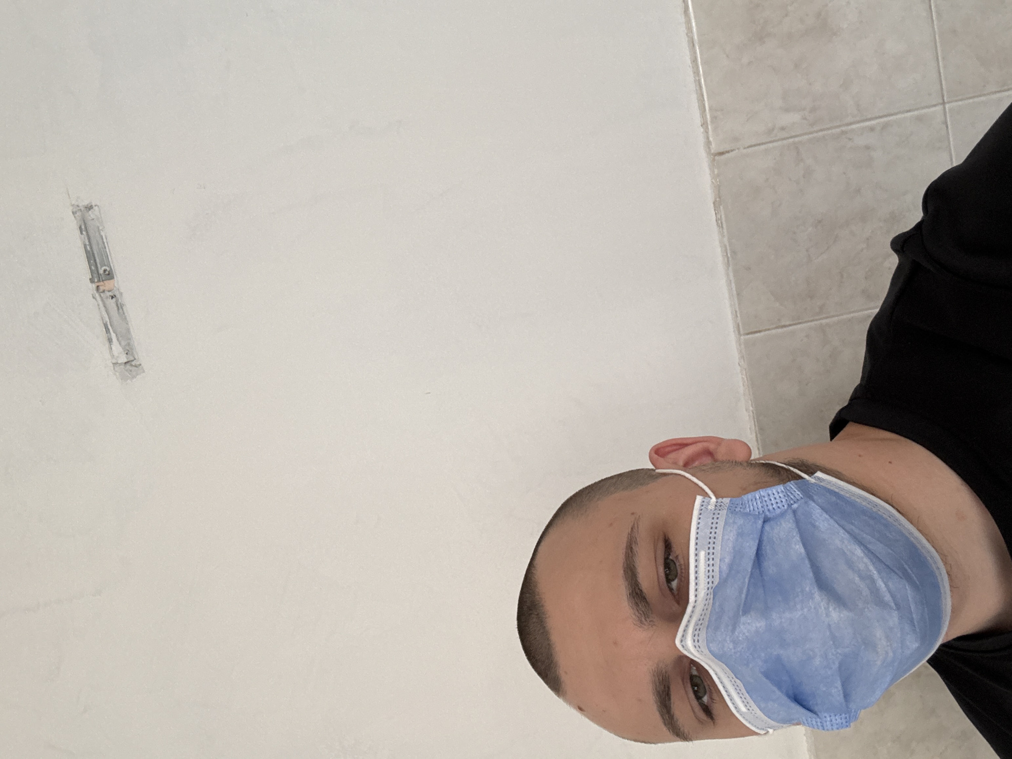 A low-angle shot shows a person wearing a blue surgical mask, viewed from below and tilted slightly to the right. The person has fair skin, short-cropped dark hair, and is wearing a black shirt. The background consists of a white wall to the left and beige tiled wall on the right. An electrical box is partially visible on the wall. It is not possible to identify the location in Stara Zagora, Bulgaria from the image.