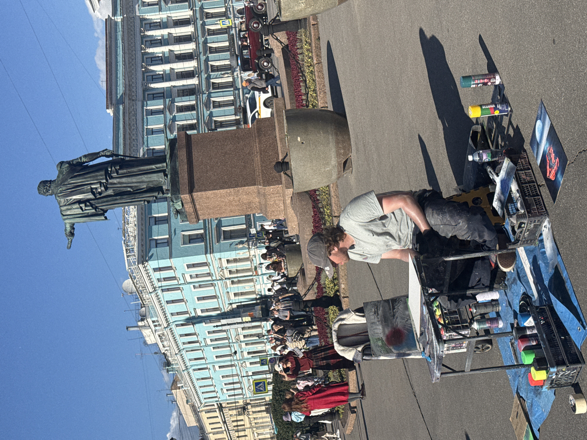In Saint Petersburg, Russia, under a clear blue sky, a street artist works outdoors. He is seated on a low stool, facing a portable easel set up on a small cart. The easel displays a partially finished artwork, with spray paint cans and art supplies arranged around it. The artist is dressed in casual clothing, including a grey t-shirt and dark pants, with a cap on his head. The scene is set in a public square with a prominent bronze statue on a pedestal in the background. Behind the statue, the facade of a light blue building with classical architectural elements is visible. Several people are walking in the background, some gathered around what appears to be another artistic display. The ground is paved with asphalt, and a blue tarp is spread out near the artist, serving as a workspace. Spray paint cans, some of them colorful, are scattered on the tarp and within plastic crates. A partially completed painting of a red car under a moonlit sky is placed on the ground in front of the artist. In the distance, vintage cars and a modern car are visible, suggesting this is a lively urban area, possibly a tourist destination.