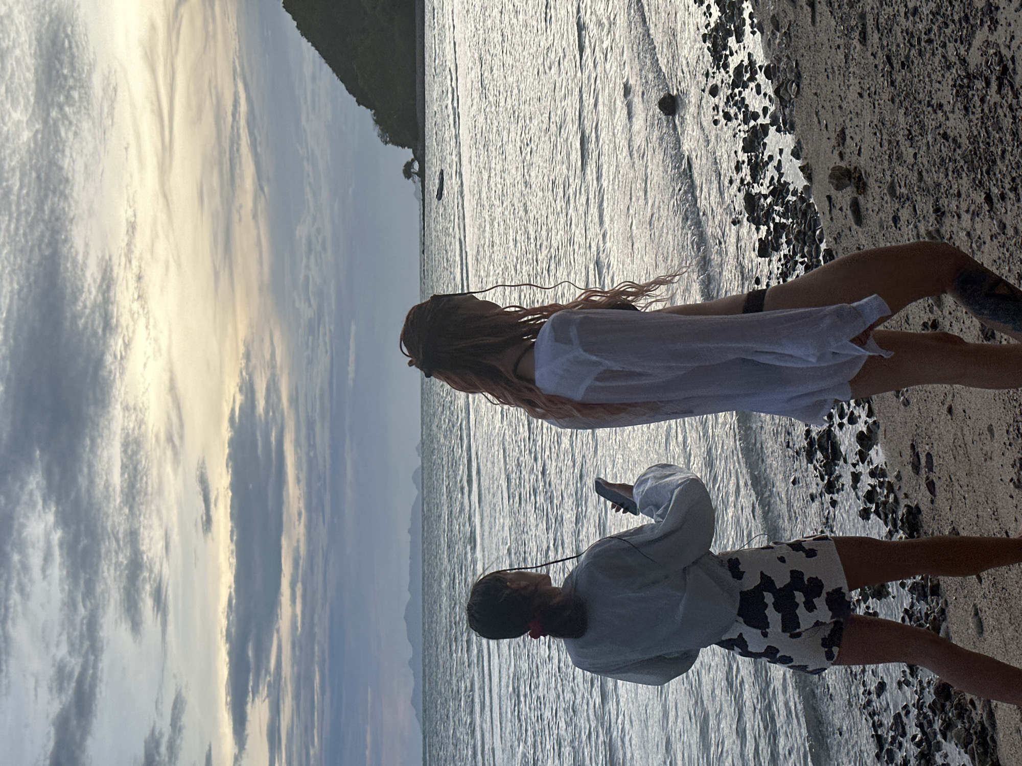 This media file depicts a scene at a beach, likely in the Ko Samui District of Thailand. Two individuals are present, viewed from behind and slightly from the side. The subject in the foreground, closer to the viewer, is wearing a light-colored long-sleeved shirt and patterned shorts. They are holding a smartphone and appear to be looking at it, possibly with headphones on. The second subject, slightly further back and taller, has long, reddish-blonde hair and is wearing a white shirt over a dark garment. Their gaze is directed towards the water.
The setting is a beach with a shoreline that is a mix of sand and pebbles. The water is calm with gentle waves reflecting the sky. In the background, there is a body of water that stretches towards a distant, hazy landmass, likely a mountainous island. The sky is filled with dramatic clouds, with patches of sunlight breaking through, suggesting it might be late afternoon or early evening, or perhaps a transition in weather. The overall mood appears serene and relaxed, with the individuals enjoying a quiet moment by the sea. No visible text is present in the image.