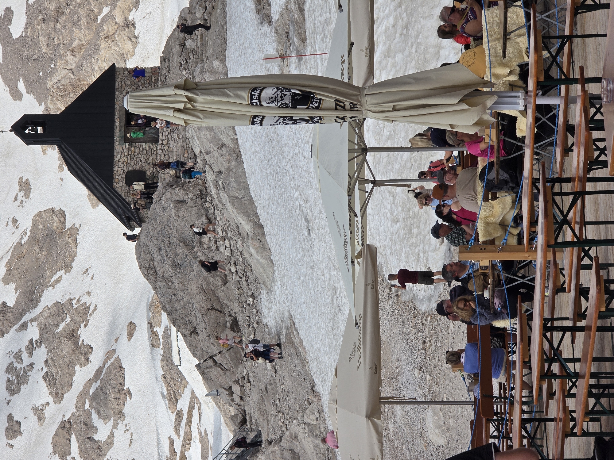 This image depicts a mountainous landscape with snow patches and rocky terrain, likely in the vicinity of Garmisch-Partenkirchen, Germany. In the background, a small stone building with a dark roof and a bell tower stands on a rocky outcrop. Several people are seen walking on the rocky paths and near the building. In the foreground, there are several outdoor seating areas with tables and benches, some covered by large umbrellas. People are seated at some of the tables, and a folded umbrella is prominently positioned in the center. The overall scene suggests a tourist or recreational area in a high-altitude environment.