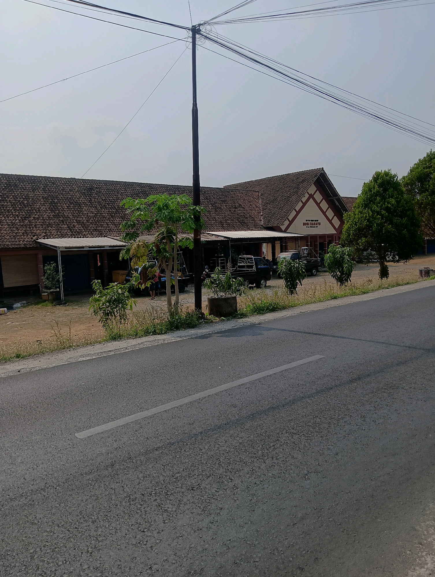 The image shows a building with a reddish-brown tiled roof, likely a business or community center, located on the side of a road in Sokaraja, Indonesia. A sign on the building reads "MUGI RAHAYU" and "BAHATU", possibly indicating the name of the establishment. Several vehicles, including pickup trucks, are parked in front of the building, with a few individuals visible. The area is surrounded by greenery, including trees and bushes. The road in the foreground is paved with asphalt and has a white line marking the lane. Utility poles and overhead wires are prominent in the sky. The overall scene depicts a typical day in a semi-urban or rural Indonesian setting.