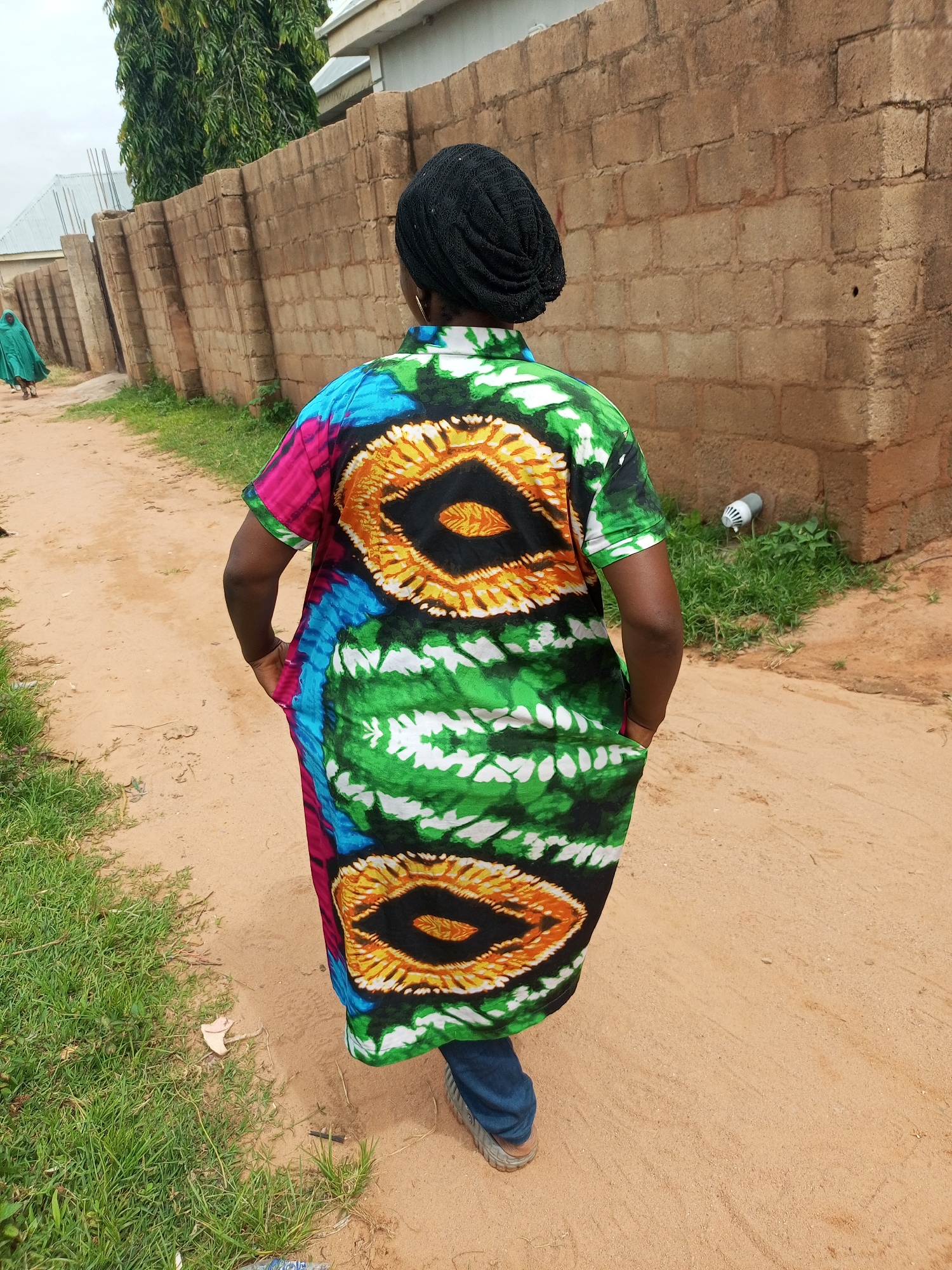 In Tempure, Nigeria, a person wearing a black headwrap and a colorful, tie-dye dress walks away from the camera on a dirt path. The dress features a striking pattern of green, white, blue, pink, and orange, with large, eye-like shapes on the back. The person is wearing blue jeans and brown shoes. To the right of the path is a brick wall, and to the left is a patch of green grass. In the distance, another person in a green garment walks along the path, and a tall tree is visible behind the wall. A white security light fixture is mounted on the wall.