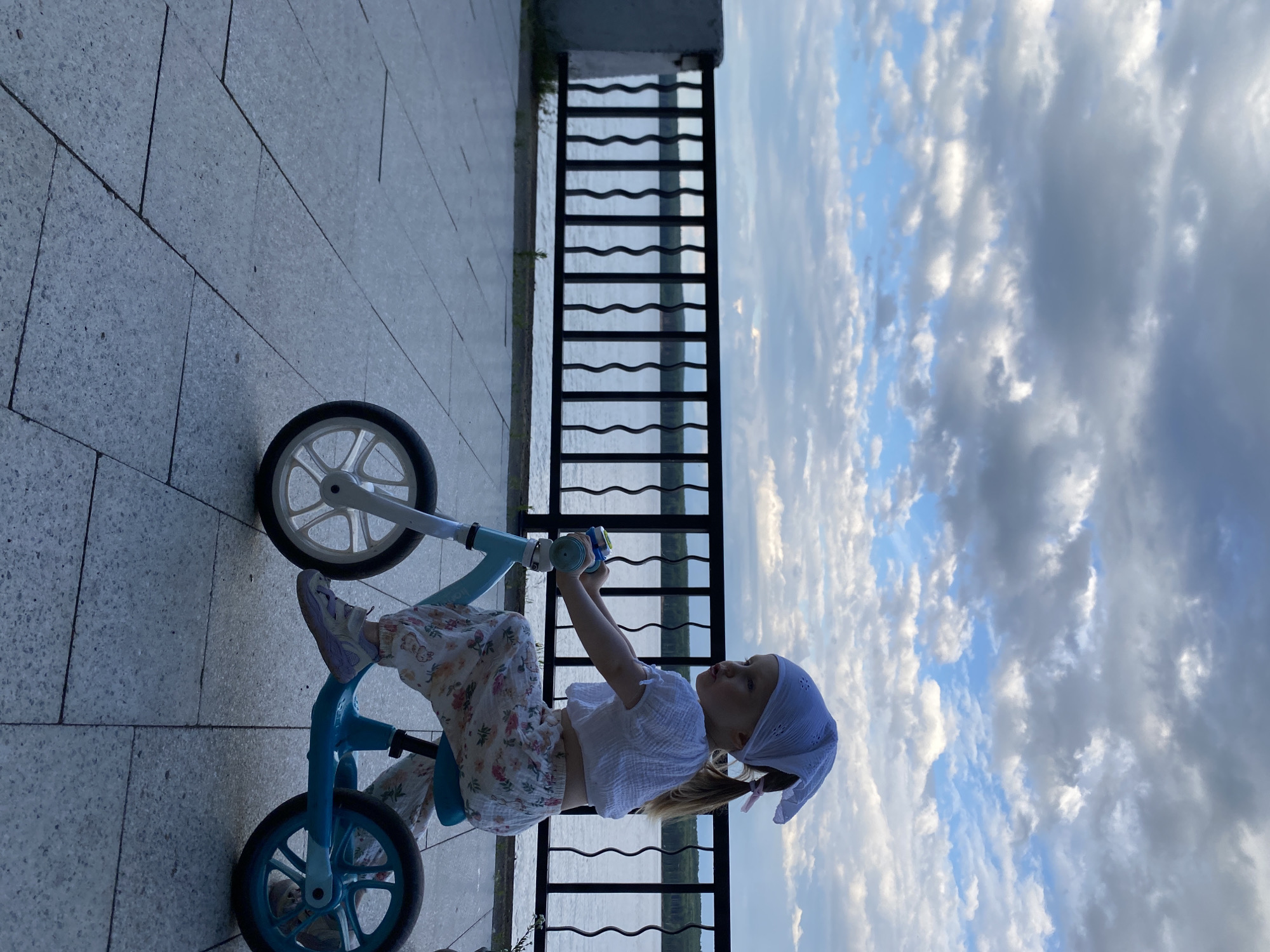 In Chaikovsky, Russia, a young girl with blonde hair tied in pigtails and wearing a white headscarf is riding a blue balance bike. She is dressed in a white cropped top and floral patterned pants, with light-colored sneakers. The girl is positioned on a paved walkway adjacent to a black metal fence. Beyond the fence lies a body of water, likely the Kama River, with a treeline visible on the opposite bank. The sky above is filled with scattered white and grey clouds against a backdrop of blue.
