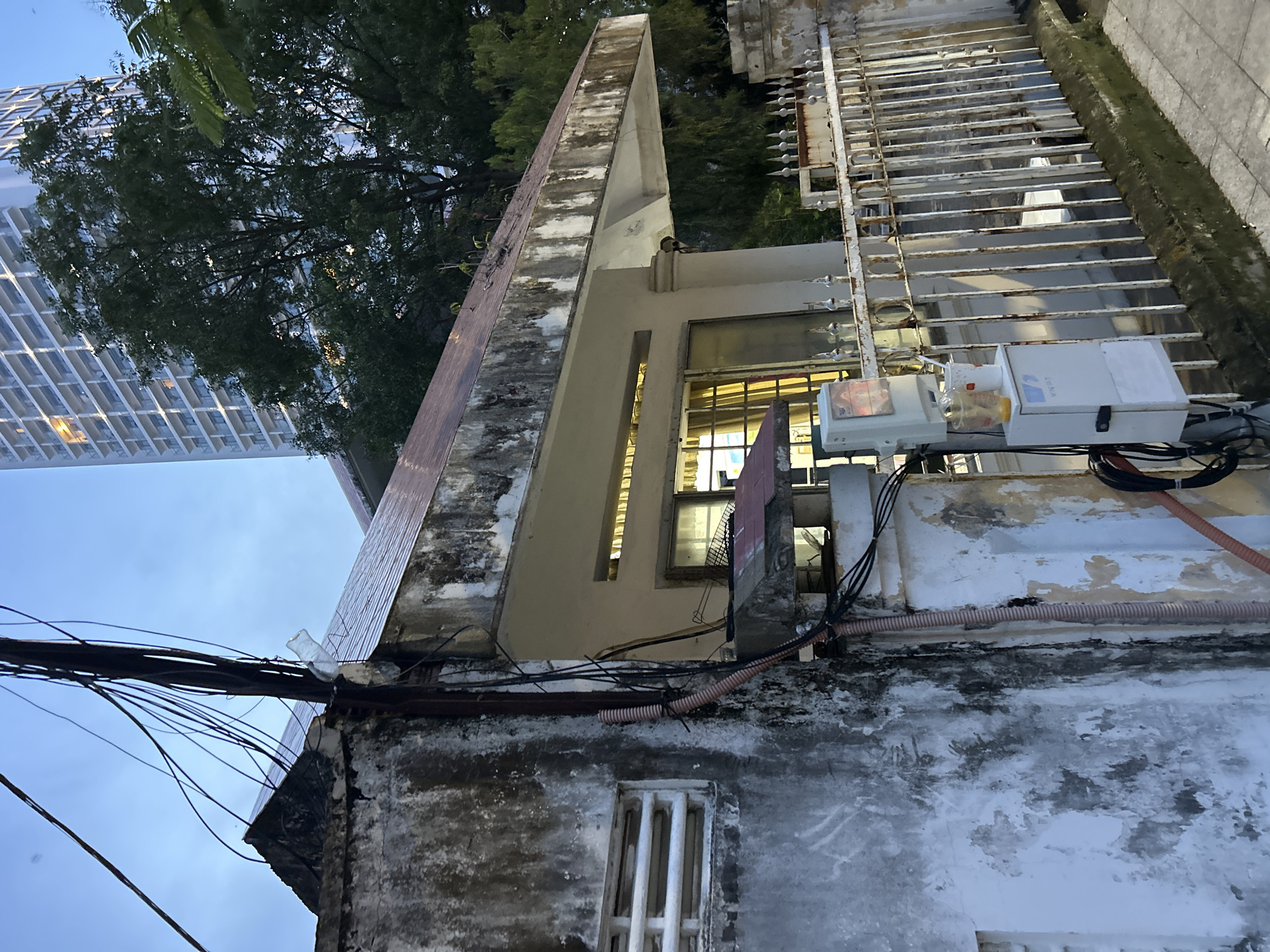The image, captured from a low angle in Nha Trang Ward, Vietnam, showcases a blend of old and new urban architecture against a clear blue sky, likely during late afternoon or early evening.
In the foreground on the right, an old, weathered building exhibits extensive wear, with peeling paint, dark stains, and grime covering its walls. A tangle of black electrical wires and orange corrugated pipes runs across its exterior, connected to several white utility boxes. One box prominently displays the brand "Loka," and another is marked with "#2." A small, barred window is visible on this lower structure. Above it, a white metal grate or fence with decorative spikes extends horizontally.
Slightly further back and to the left, another building section features a distinctive, sharply angled concrete roofline. A window on this part of the building has a metal grate, and an interior light is visible through it. A small bird is perched on the concrete edge of this structure.
To the far left and in the background, a modern, tall skyscraper with a grid-like facade of windows rises into the sky, contrasting sharply with the aged structures. Dense green trees partially obscure the base of the skyscraper and fill the space between the buildings. A clear plastic bottle is visible caught within the complex network of wires near the old building's roof. No human activity is depicted in the scene.