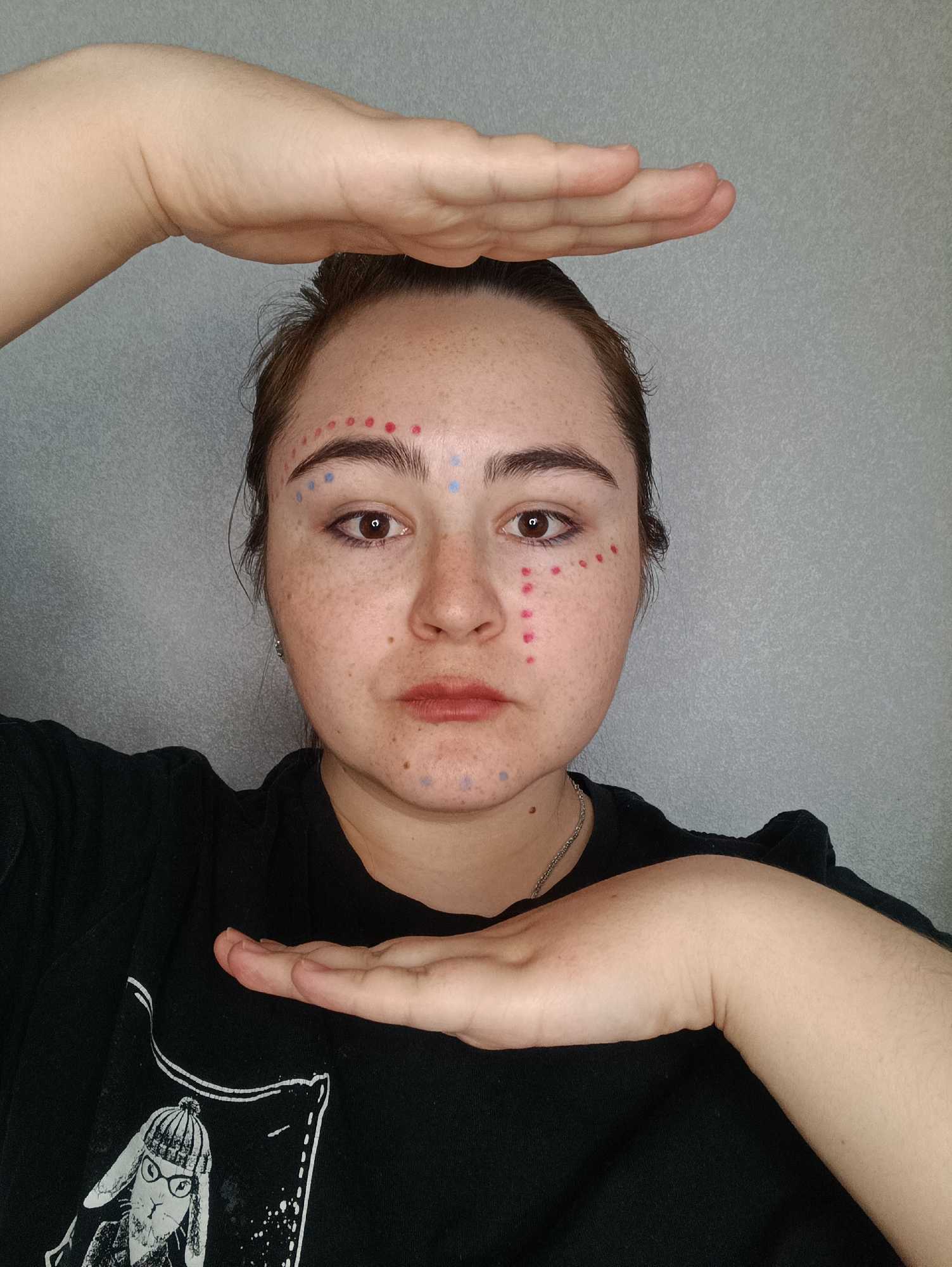 A fair-skinned person with freckles faces the camera, positioned centrally in the frame. Red and blue dots are applied to the person's face in a pattern. The individual is wearing a black t-shirt with a white graphic of a rabbit wearing glasses and a beanie. The person's hands are positioned above and below their face, palms facing inward as if framing their face. The background is a plain gray wall. There is no information to suggest that this image was taken in the city of Noviy, Russia.