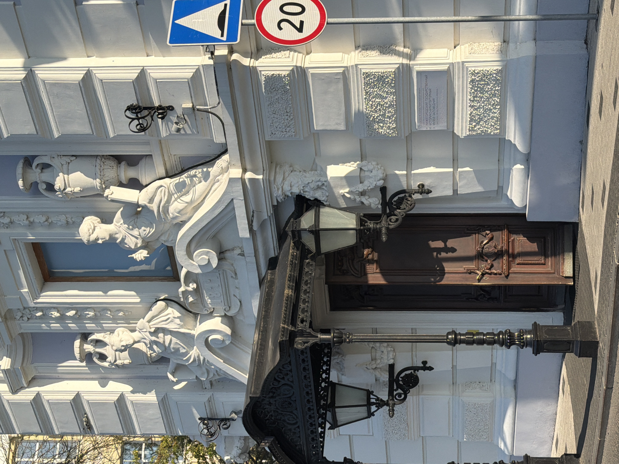The image shows the facade of the Nizhny Novgorod State Museum of Architecture and Landscape, located in Nizhny Novgorod, Russia. The building is white with classical architectural elements, including ornate carvings, statues, and decorative urns. A dark wrought-iron awning with two hanging lanterns covers the entrance, which features a large, dark wooden door with intricate carvings. A street lamp stands in front of the entrance. Traffic signs indicating a speed limit of 20 and a speed bump are visible on the right side of the image. A plaque with Russian text is affixed to the building below the signs. The scene is bathed in sunlight, casting long shadows.