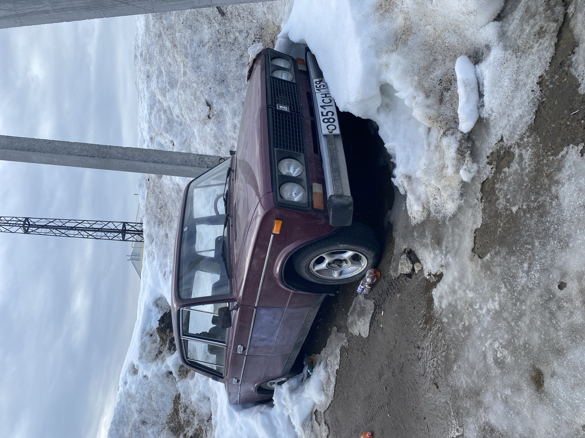 The image displays a maroon VAZ-2105-series sedan, registered with Russian license plate O851CH 159 (Perm Krai region), parked partially submerged in dirty, melting snowdrifts on what appears to be a wet asphalt or dirt surface. The scene is set in Chaikovsky, Perm Krai, Russia, under an overcast sky.
The vehicle is positioned with its front end facing slightly right, directly adjacent to a large accumulation of snow and ice. The driver's side rear is particularly enveloped by snow. The car's paintwork exhibits dirt and a noticeable faded area on the driver's side front door. The driver's side front wheel features a silver multi-spoke alloy rim. The passenger-side headlight cluster is partially obscured by snow.
The ground around the vehicle is a mix of wet, dark soil/asphalt and patches of melting ice, with visible tire tracks and puddles. Several pieces of refuse, including multiple plastic bottles containing liquid and other small debris, are scattered on the ground near the car.
In the background, a tall, dark metal lattice tower is visible to the left of the vehicle. To the right, two large, grey concrete utility poles stand vertically, partially framing the scene. A light-colored building is discernable in the distant background, beyond the snow piles. No individuals are present in the image.