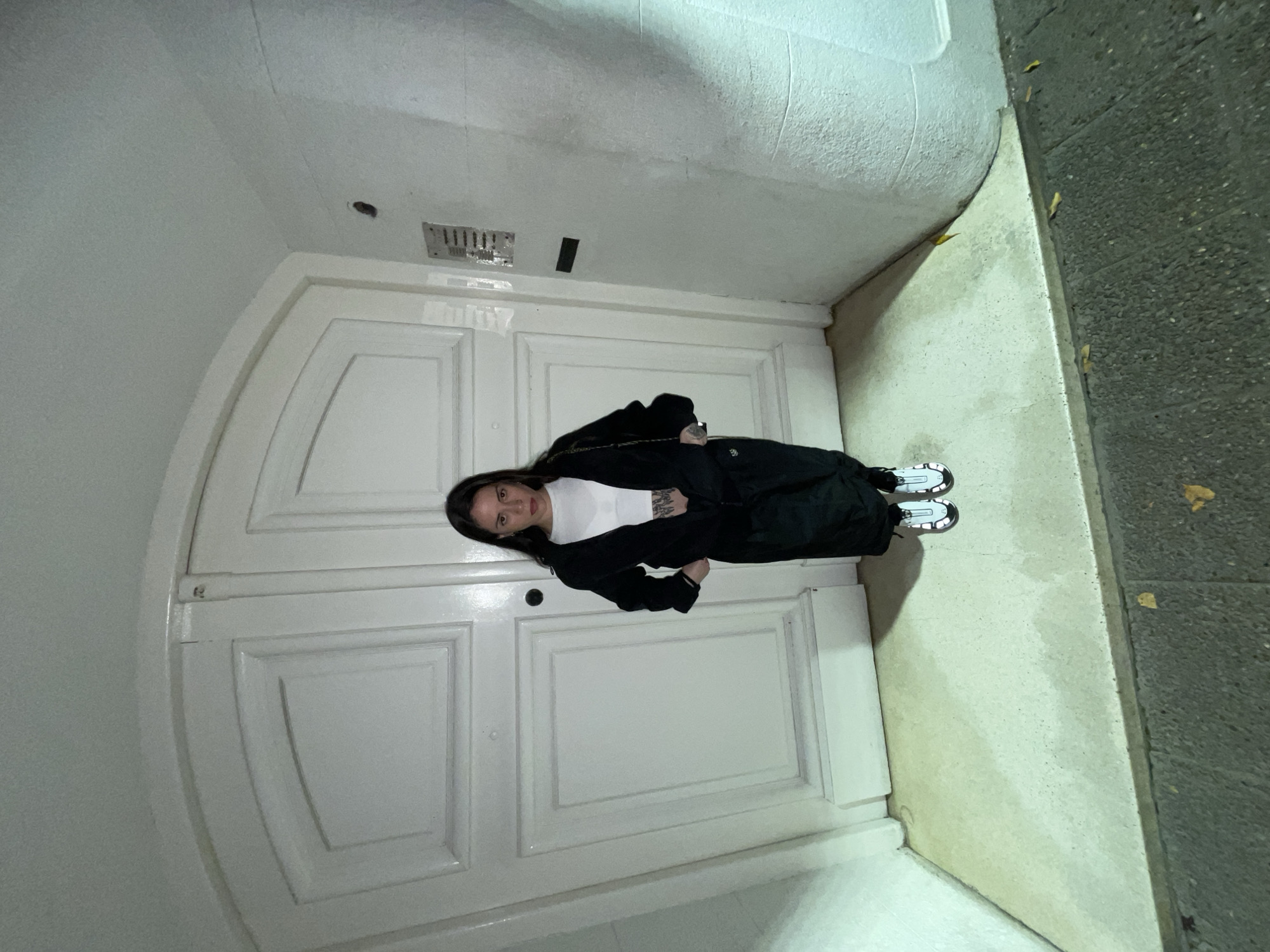 A woman is standing in front of a large white arched doorway. She is wearing a white shirt, black pants, and a black jacket. The doorway is framed by a white wall that curves into the ceiling. To the right of the doorway is a sidewalk or street made of grey stone, with a few fallen leaves scattered on it. Based on the architectural style of the doorway and the surrounding structures, this location may be in the city of Buenos Aires, Argentina.