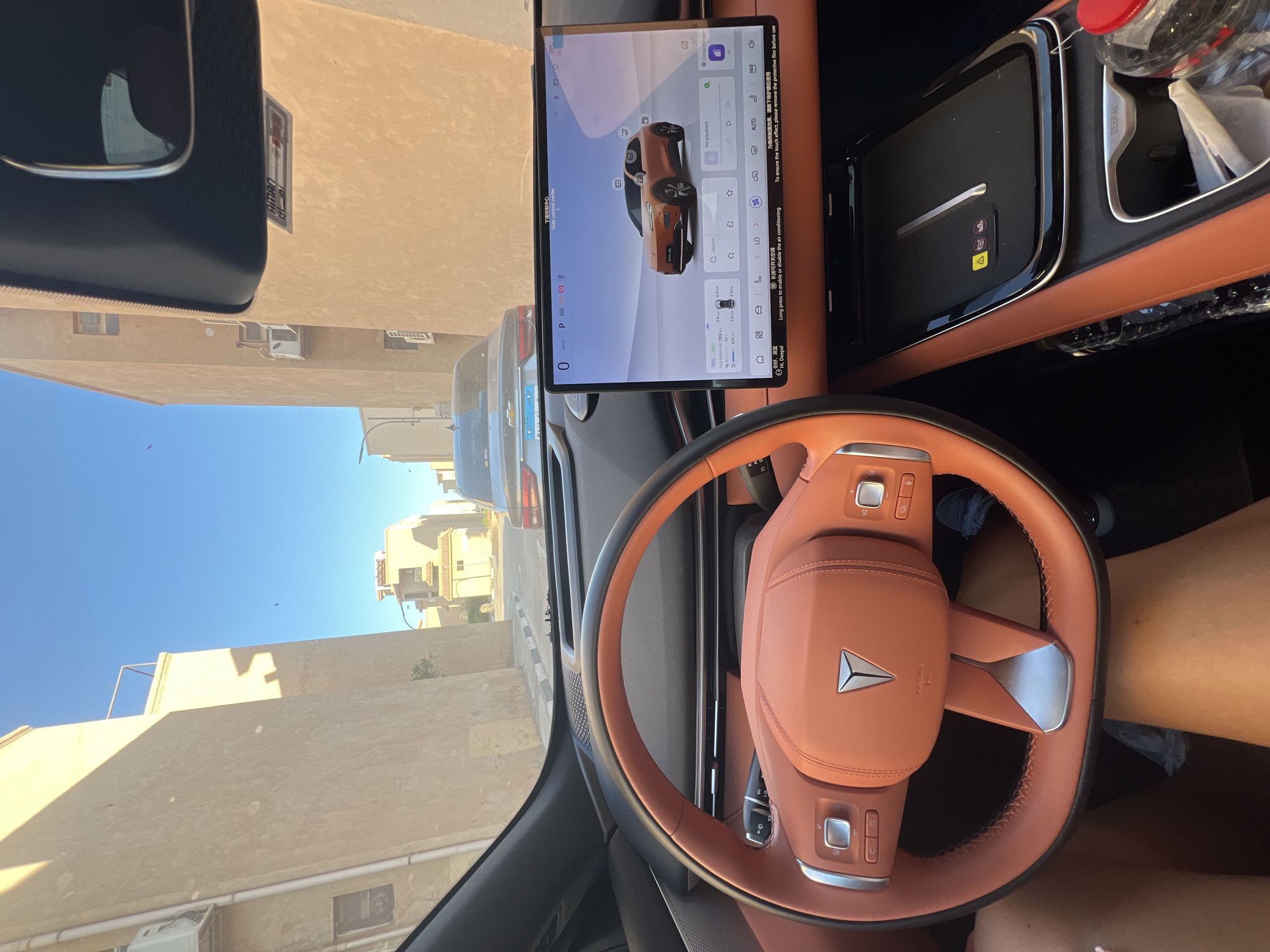 The image shows the interior of a modern car, looking forward through the windshield. The car is parked or driving on a narrow street lined with two-story buildings made of light-colored stucco. The sky is clear and blue.
In the foreground, the orange leather steering wheel is prominent. To the right of the steering wheel is a large central touchscreen display showing a car interface with an orange car illustration. Below the screen is a black wireless charging pad with a warning symbol and two smaller icons.
In the background, two cars are visible on the street. The car directly in front has a license plate that appears to be Egyptian. The street surface is asphalt with a sidewalk demarcated by a curb and a white and black patterned border. The buildings have visible air conditioning units mounted on their walls.
The overall impression is that of a sunny day in an urban environment, likely a residential or commercial area, with a focus on the car's interior technology.