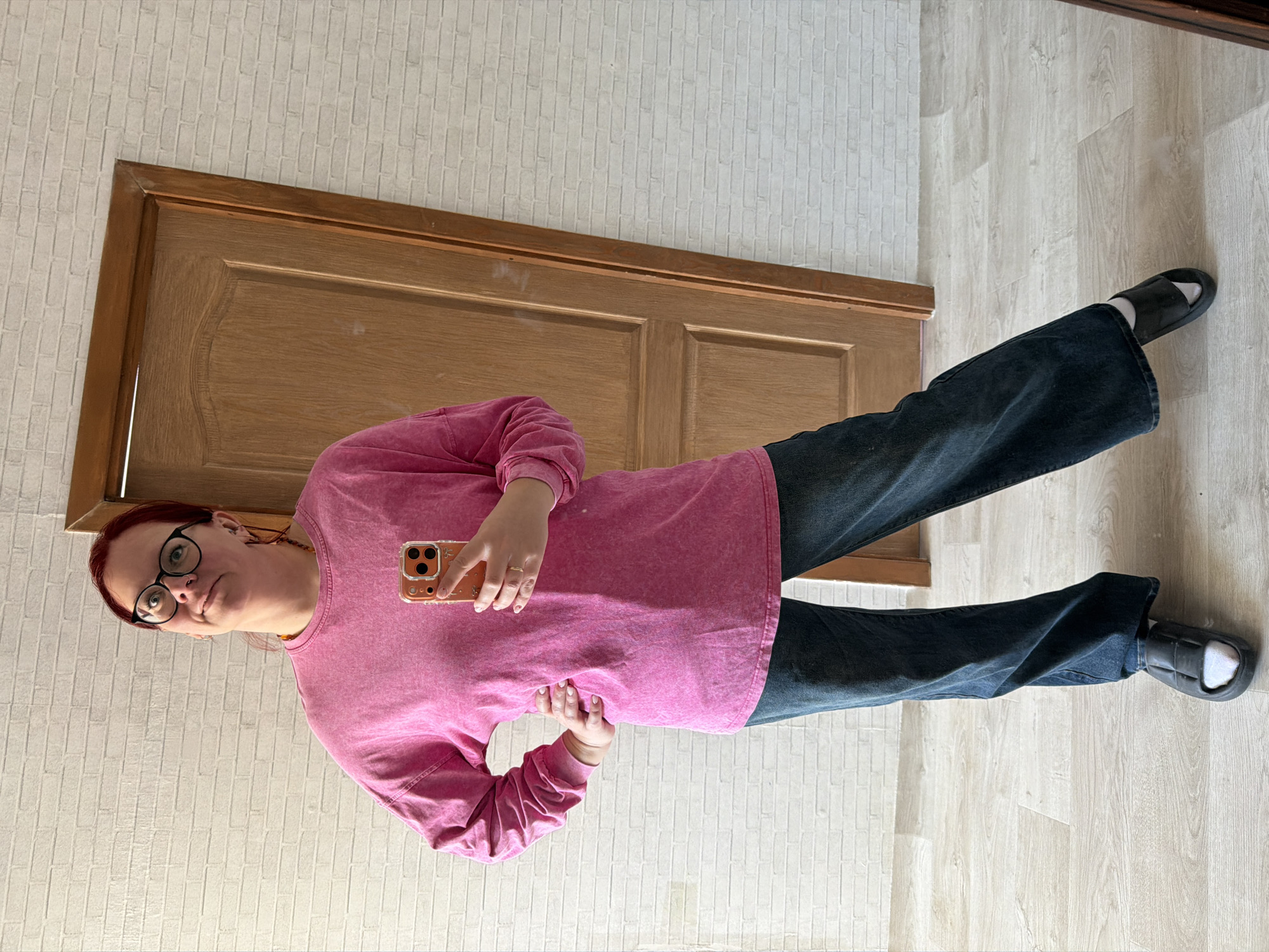 An indoor full-body image depicts an adult individual standing in what appears to be a residential or commercial space in Chaikovsky, Russia. The person has reddish hair, wears black-framed glasses, a nose stud, and an amber-colored beaded necklace. They are dressed in a long-sleeved, fuchsia-pink, acid-washed shirt, dark wide-leg jeans, and black padded slide sandals worn over white socks. A thin gold-colored ring is visible on the ring finger of their right hand. The individual holds an orange-colored smartphone with a clear case in their right hand, positioned as if taking a mirror selfie. Their left hand is placed on their hip, and their gaze is directed upwards and to the left with a slight facial expression. The background features a light-colored wall with a textured pattern resembling thin bricks. To the right of the individual, a light brown wooden door with paneling details is partially visible. The floor consists of light wood-grain laminate planks arranged in an irregular pattern.