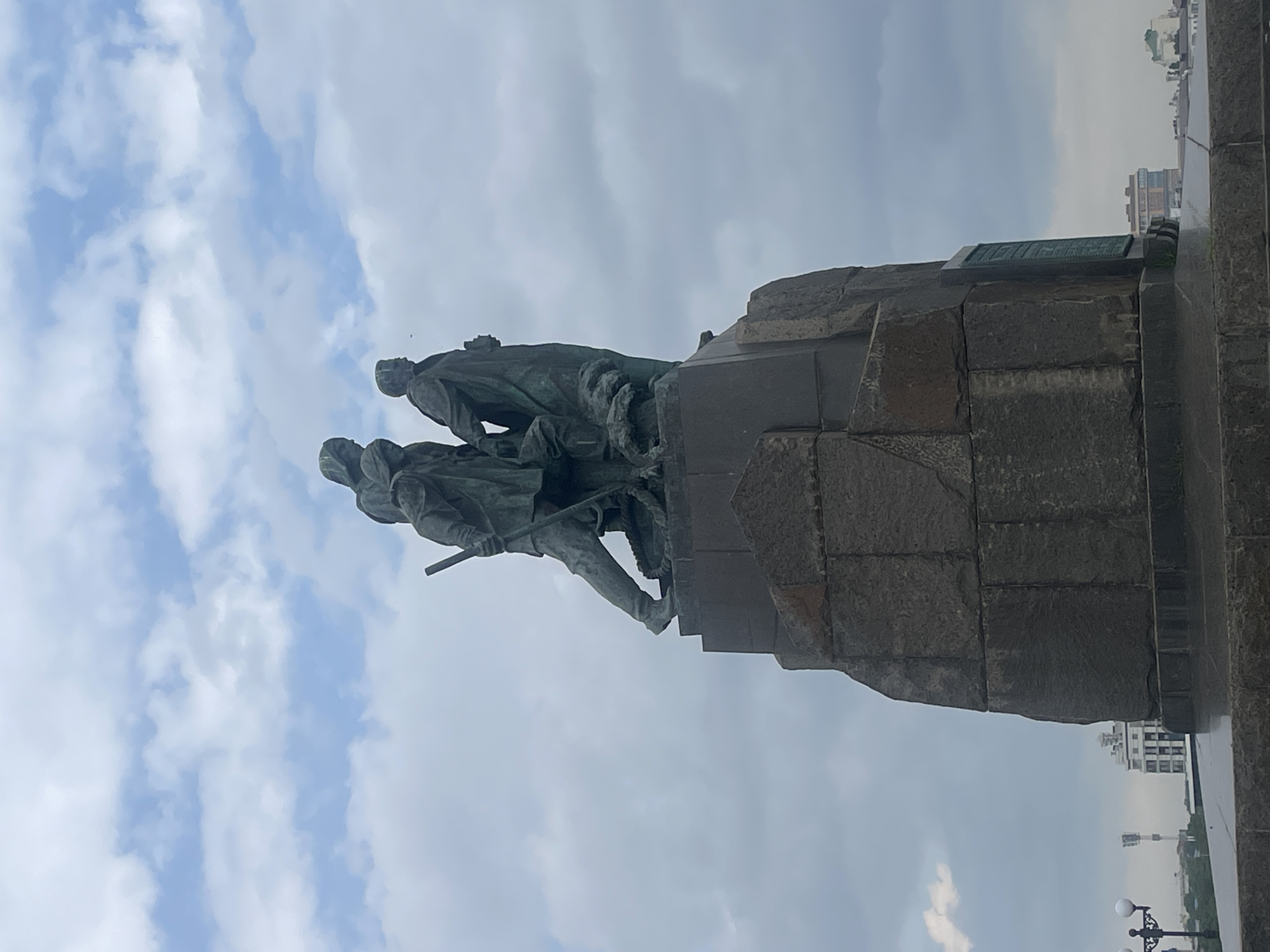 This is a vertical, eye-level, medium shot of a monument depicting three figures standing on a large stone pedestal. The monument is made of bronze and appears to be weathered. The figures are cloaked and appear to be walking together. One figure on the left is holding a staff. The pedestal is made of rough-hewn stones. In the background, a cloudy sky with patches of blue is visible, along with distant buildings and a lamppost.
The monument is the "Monument to the Sailors and Defenders of Novorossiysk" in the city of Novorossiysk, Russia. It commemorates the sailors and defenders who fought in the Russian Civil War and World War II. The monument stands on a hill overlooking the Black Sea. The statue was designed by sculptor Ivan Shadr and architect Lev Rudnev and was erected in 1948.