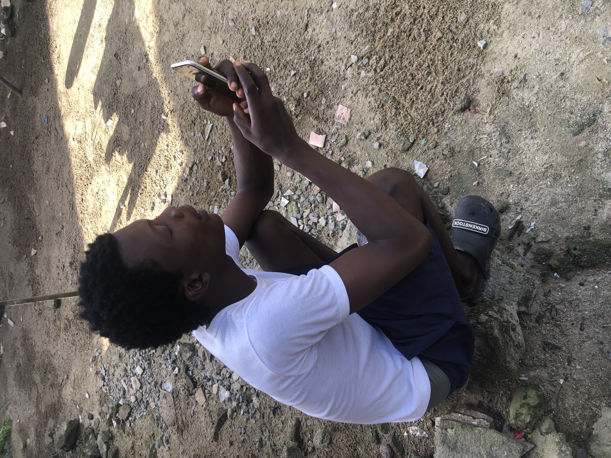 A young Black male is squatting on sandy ground with scattered rocks and debris, holding a smartphone. He is wearing a white t-shirt and dark shorts, with a greyish-green band visible at the waistband. His feet are clad in black slides with "BIRKENSTOCK" written on them. The background consists of more sandy ground with shadows cast by an unseen object, suggesting an outdoor, possibly unpaved area. The scene appears to be in Ajah, Nigeria, characterized by the natural lighting and the informal outdoor setting.