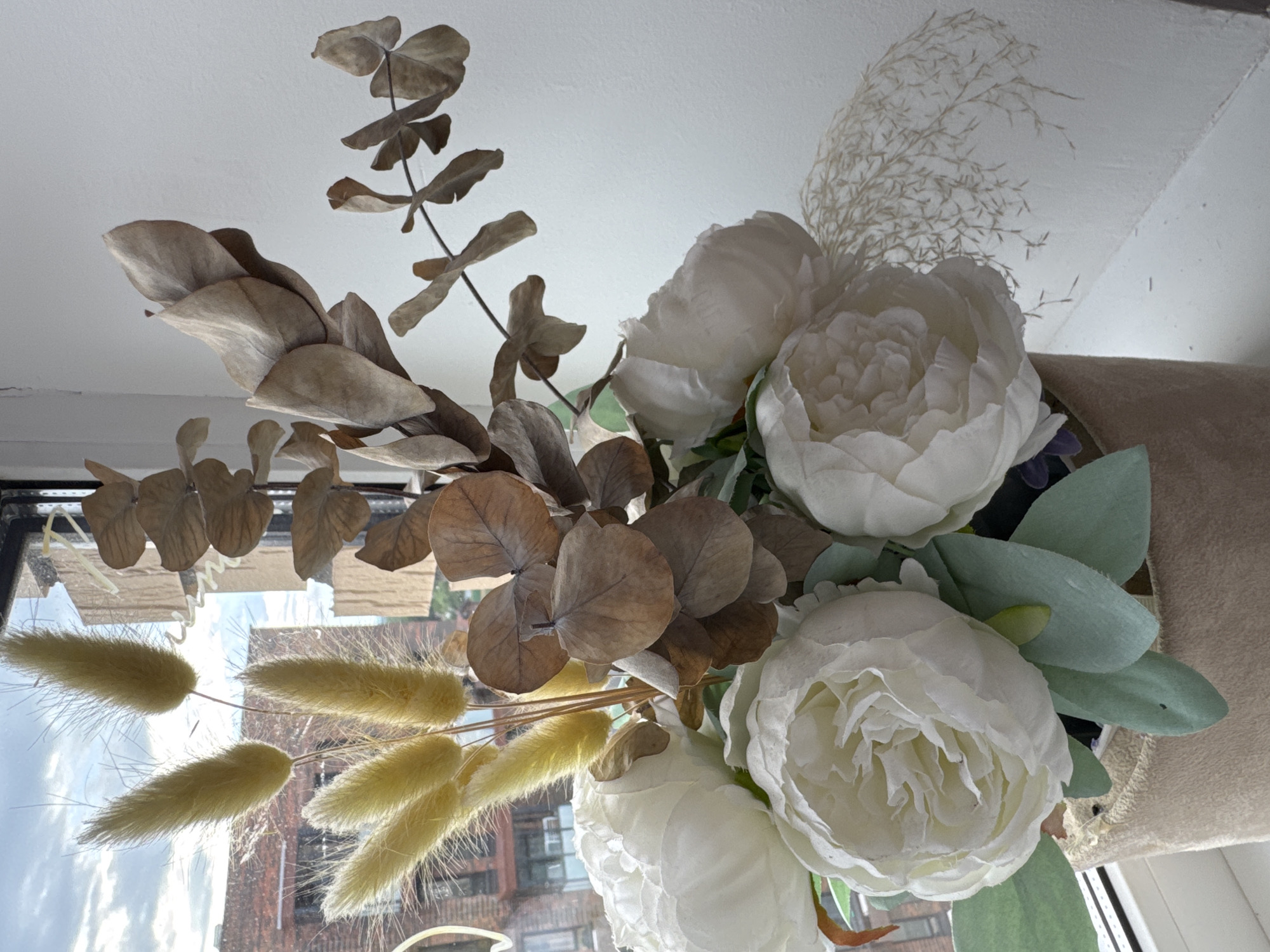 A still life photograph shows a bouquet of artificial flowers, including white peonies and dried eucalyptus leaves, arranged in a pot. The bouquet is positioned in front of a window, with the cloudy sky and brick buildings visible through the glass. The lighting suggests it may be daytime. The overall mood is neutral, with a focus on the textures and colors of the floral arrangement. There is no visible text in the image.