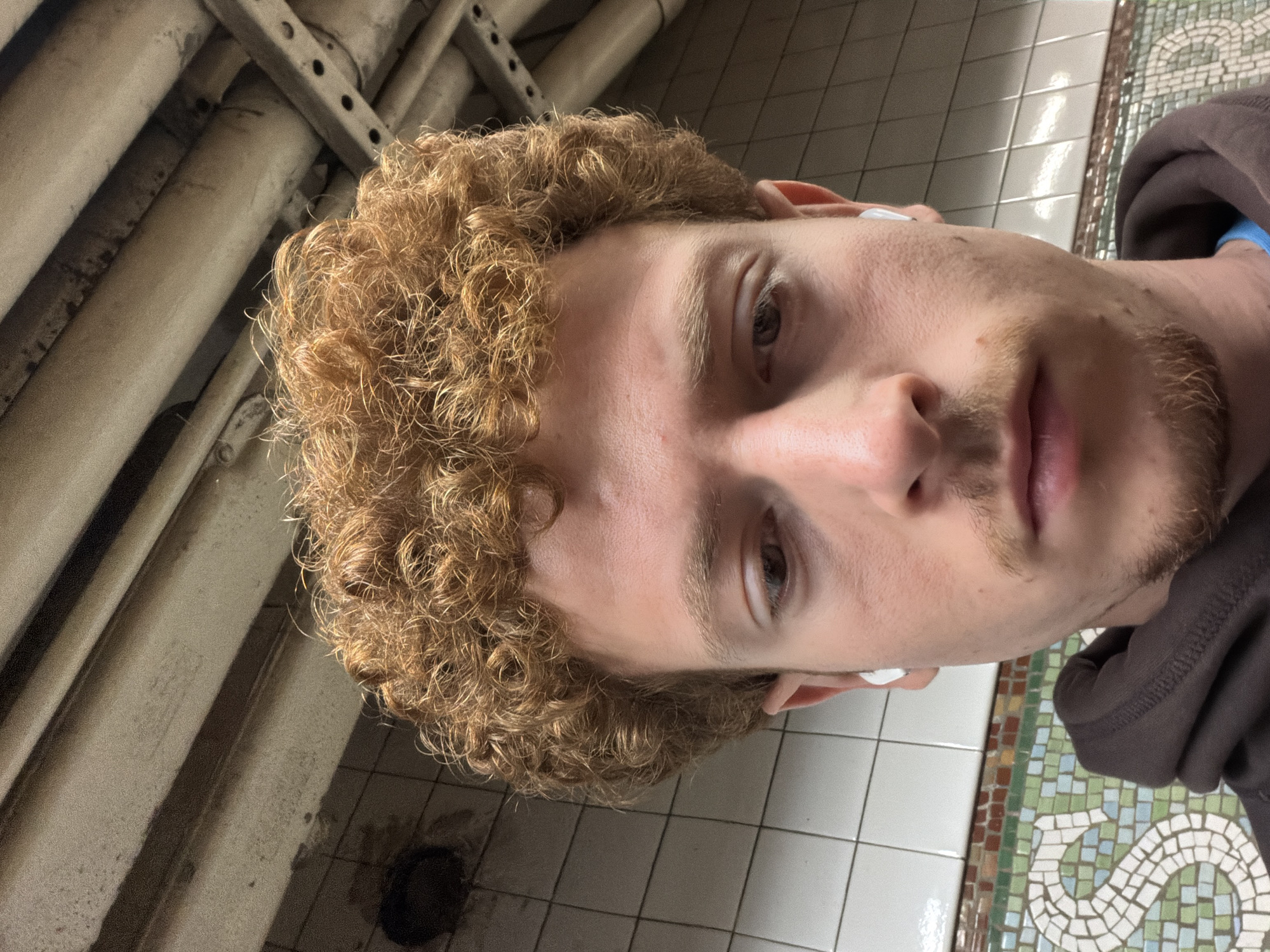 The image is a close-up of a man with curly blonde hair. He has fair skin and light eyes. He is wearing a wireless earbud in his right ear. He has a thin beard. The man appears to be in a subway station in New York City. The background includes subway pipes and tiled walls. The floor is also tiled with a mosaic pattern.