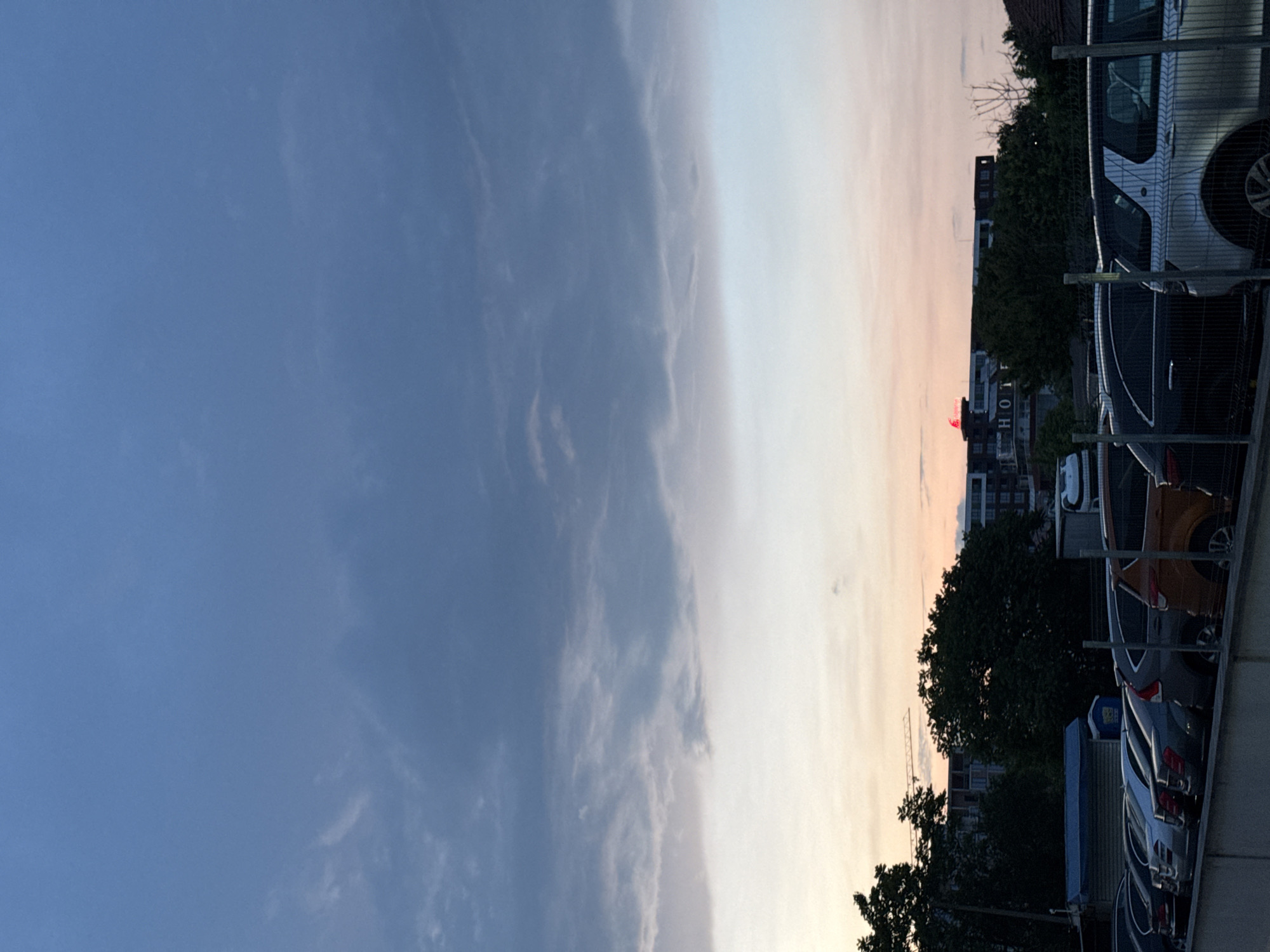 The image captures an urban landscape at dusk or sunset in Burgas, Bulgaria. The sky dominates the upper portion, displaying a vibrant gradient from a deep, clear blue on the left to softer, warmer hues of pale orange and pink on the right, characteristic of twilight. Scattered, wispy clouds are visible across the sky, more distinct in the bluer areas.
Along the lower half of the image, a diverse skyline of buildings and trees is silhouetted against the luminous sky. A prominent multi-story building features the word "HOTEL" written vertically down its side. On a rooftop slightly above and to the left of the "HOTEL" sign, a small, bright red structure, possibly an illuminated sign or an antenna, stands out. Further in the background, a construction crane is visible, rising above the distant buildings and trees.
Dark green trees are interspersed among the urban structures, their foliage appearing dense against the fading light. In the foreground, particularly on the right side of the image, there is a parking area containing several parked cars of various colors, including silver, dark grey, and orange. These vehicles are partially obscured by a wire fence or barrier that runs across the foreground. The overall scene depicts a serene urban transition from day to night.