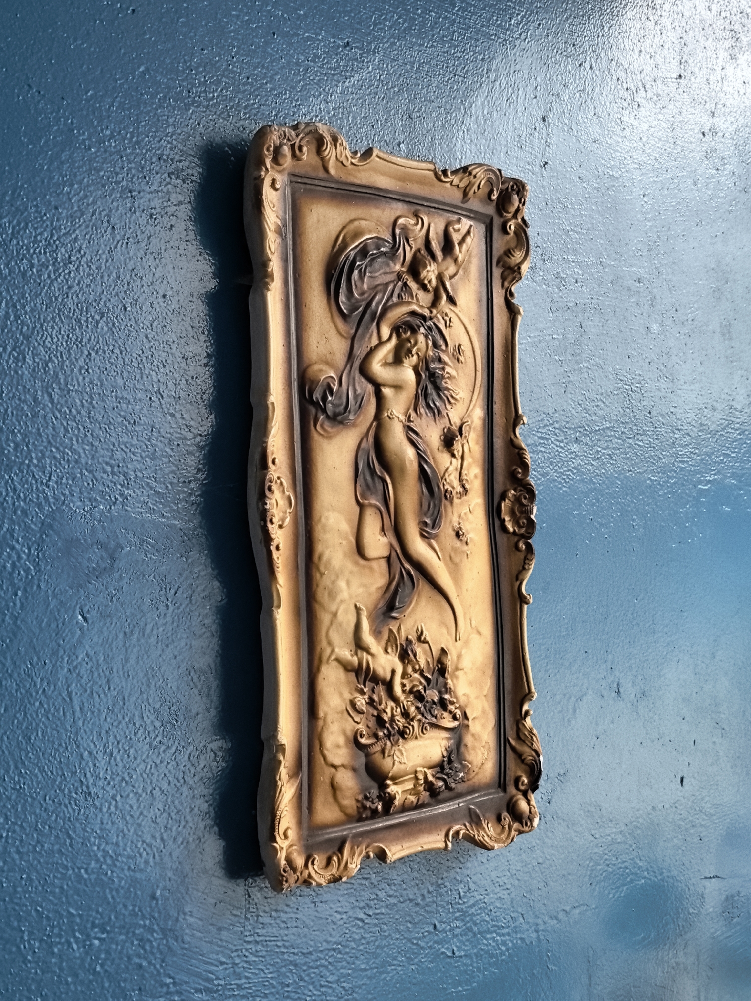 An ornate, framed relief carving of a classical scene featuring a nude female figure with flowing drapery is mounted on a textured, blue wall. The carving depicts the woman in a dynamic pose, with several cherubic figures and floral elements surrounding her. The frame is decorative, with elaborate scrollwork and details. The lighting casts a shadow on the wall behind the artwork, indicating its placement is likely indoors and not in direct sunlight. The overall aesthetic suggests an antique or vintage decorative piece.