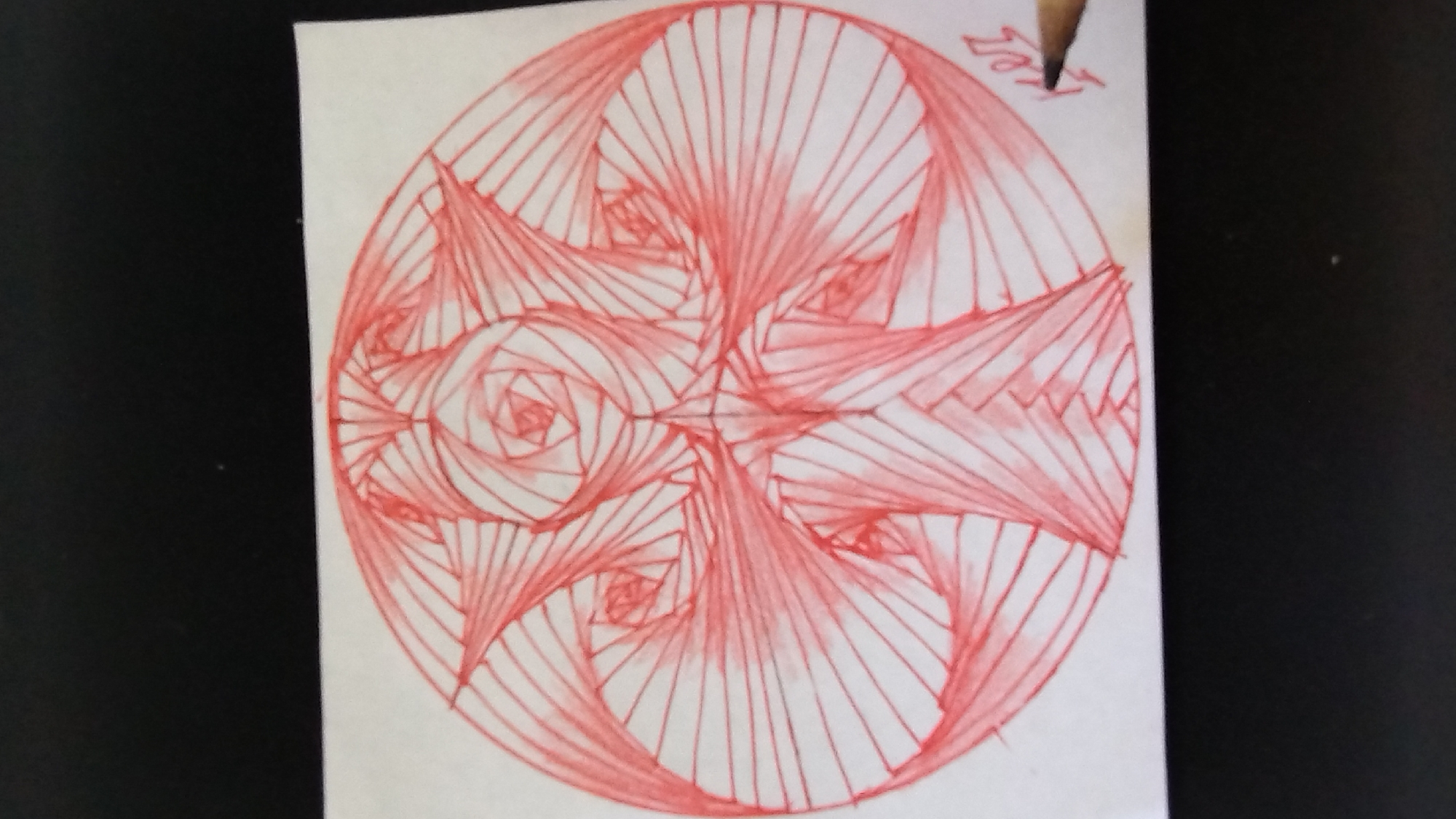 This is a close-up, overhead shot of a piece of white paper with a drawing on it. The drawing is done in red ink and appears to be a geometric, radial design. The design is composed of many thin, parallel lines that converge at a central point. These lines create a swirling, fan-like pattern that extends outwards towards the edges of the paper. Within the larger radial pattern, there are smaller, spiraling elements that resemble stylized roses or geometric knots.
In the upper right corner of the paper, there is some text written in red ink. The text appears to be a name or signature and reads "Jel" written vertically, with the "J" at the top and the "l" at the bottom. A pencil tip is visible in the upper right corner, suggesting the drawing may have been recently made or the signature added. The paper itself is slightly creased and worn at the edges, indicating it might be a sketch or a page from a notebook. The background is dark and out of focus, likely a table or surface. The overall impression is that of an abstract, artistic doodle created with precision and care.