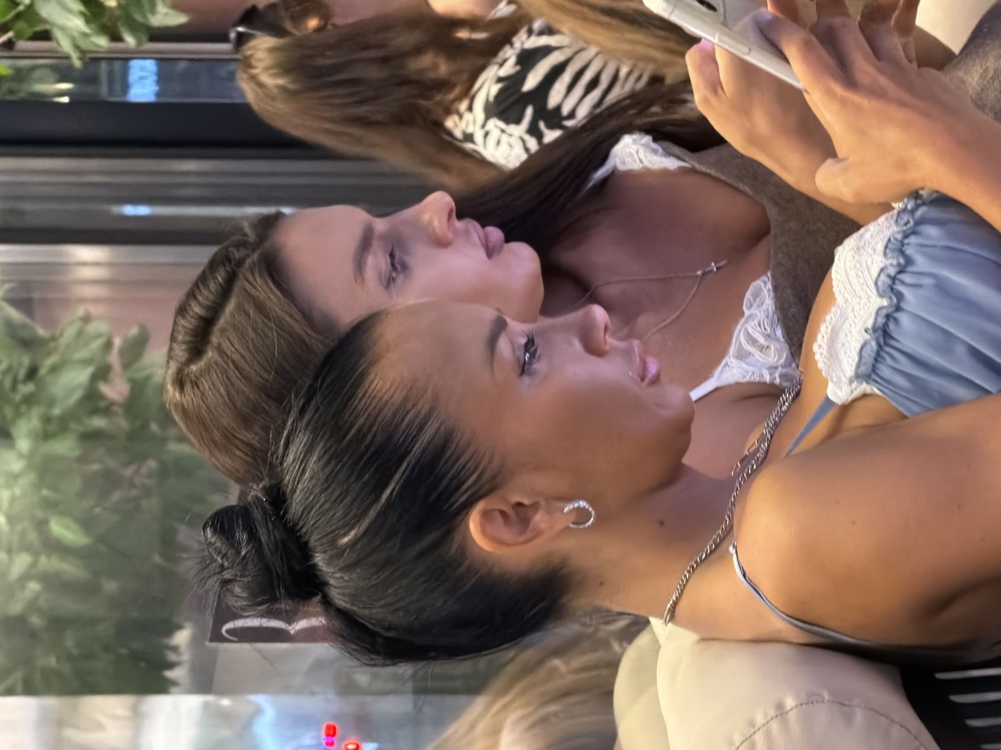 The image shows a medium shot of two women sitting close together, possibly in an indoor cafe or restaurant. The woman in the foreground on the left has her dark hair pulled up in a bun and is wearing a light blue dress with white lace trim. She has a silver hoop earring and a silver chain necklace with a cross pendant. The woman next to her on the right has long brown hair and is wearing a brown top with white lace trim. Both women are looking down at their phones. The background is slightly blurred but shows greenery and some indoor lighting, suggesting a public indoor space in Novaya Usman', Russia.