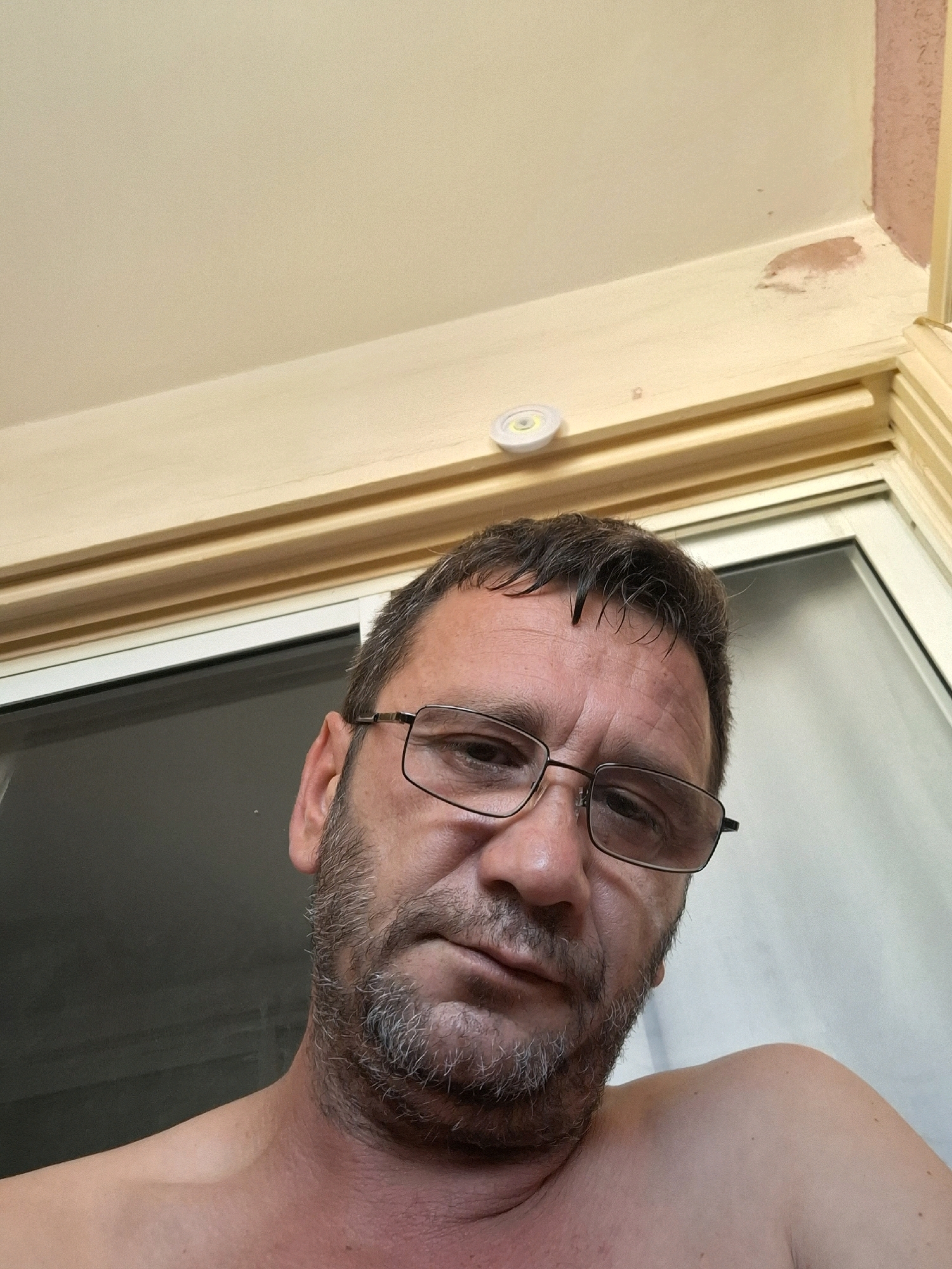 The image is a close-up, low-angle selfie of a man wearing glasses, with a beard and short hair. He is looking directly at the camera. The background consists of a beige ceiling with some light damage, a wooden window frame, and a window with a white curtain. There is a small white disc-shaped object attached to the ceiling near the corner. The man appears to be indoors, possibly on a balcony or near a window. The scene does not contain any specific identifiable landmarks or features that would indicate a location in Yerocham, Israel.