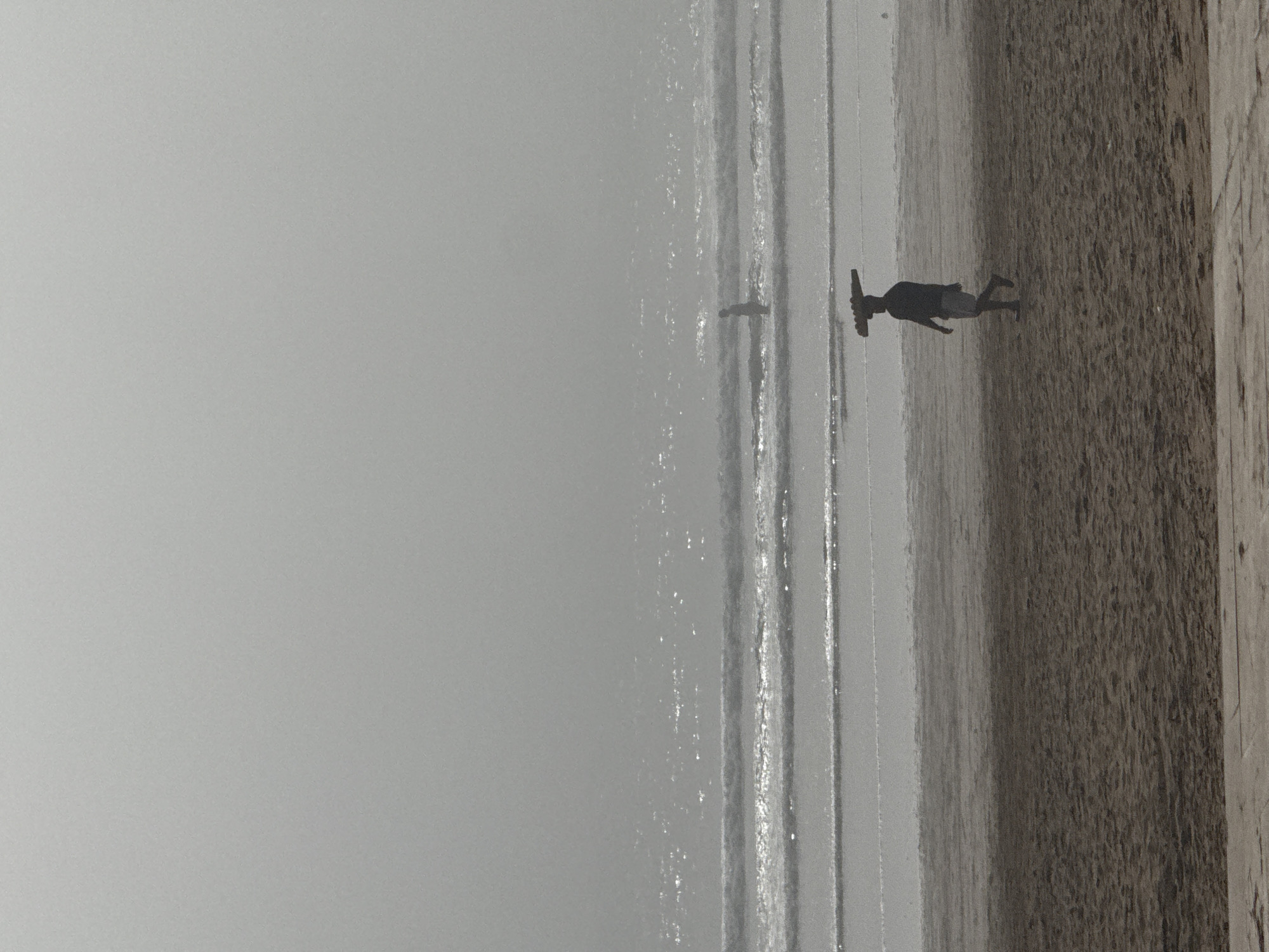 The image shows a beach scene in Agadir, Morocco. The sky is overcast, creating a muted gray tone across the scene. The sandy beach, darker in color, takes up the right portion of the image, while the ocean occupies the central part with visible waves moving toward the shore. A person is walking along the shoreline near the edge of the water. Another individual can be seen in the water.