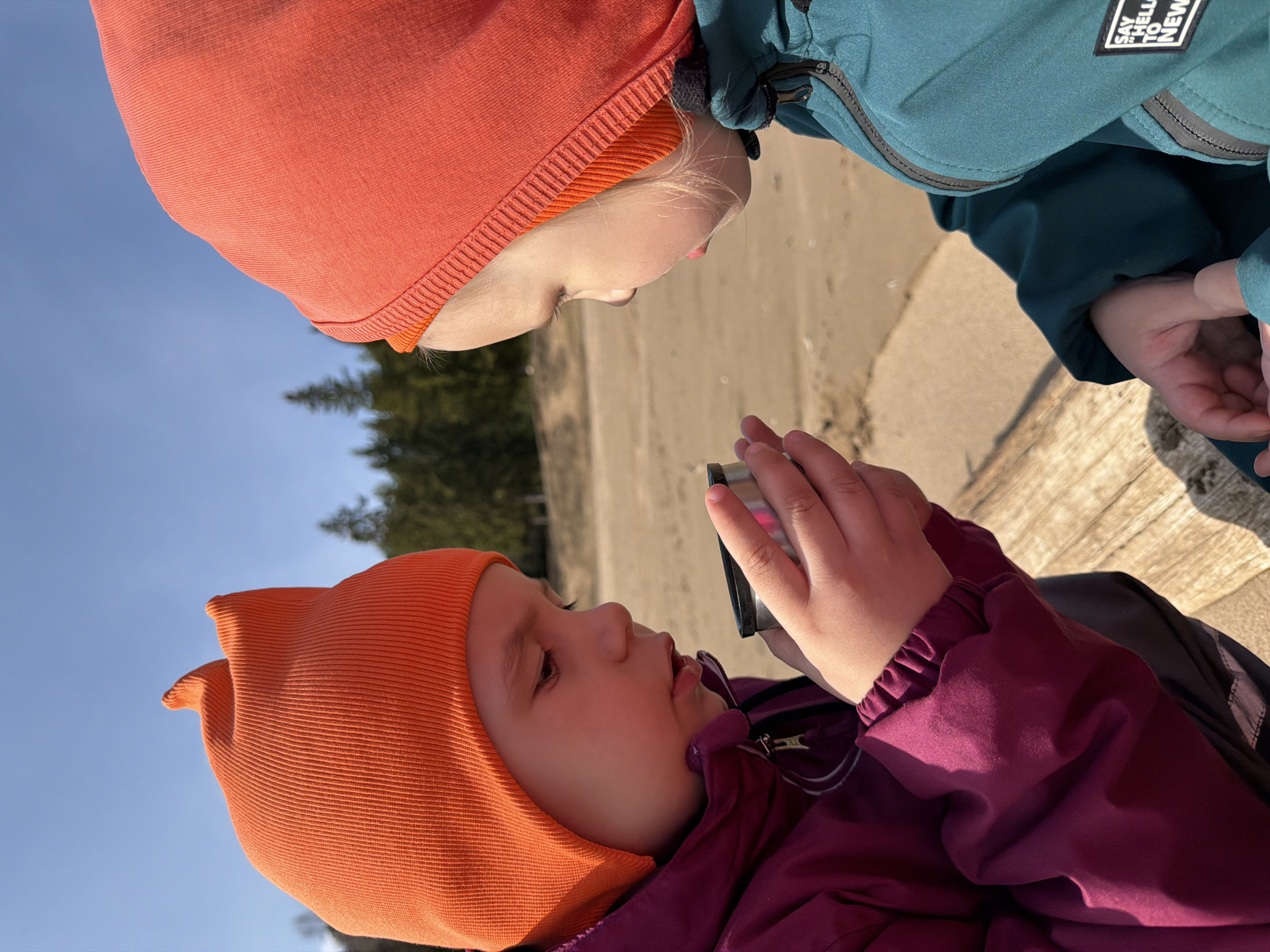 The image, rotated 90 degrees clockwise, shows two young children engaging in an outdoor setting in Chaikovsky, Russia, on a bright, sunny day.
The child on the left wears an orange knitted balaclava or hat and a purple/maroon jacket. They are holding a small, metallic container or bottle, possibly a mini-thermos, with both hands and looking downwards and to the right at the object.
The second child, positioned above and to the right, wears a similar orange knitted balaclava or hat and a teal/dark green jacket. On the right sleeve of this jacket, a black and white patch is visible, clearly displaying the text "SAY HELLO TO NEW". This child is looking directly at the first child and the object, with a soft, inquisitive expression. Both children have fair hair peeking out from under their hats.
The background features a clear, bright blue sky in the upper left corner and a sandy or gravelly ground surface that stretches into the mid-ground. Dark green, coniferous trees are visible in the distant background. The bright sunlight suggests it is daytime, possibly morning or afternoon, with no clouds in the sky. The children are in close proximity, suggesting a shared moment of curiosity or interaction with the object one of them is holding.
