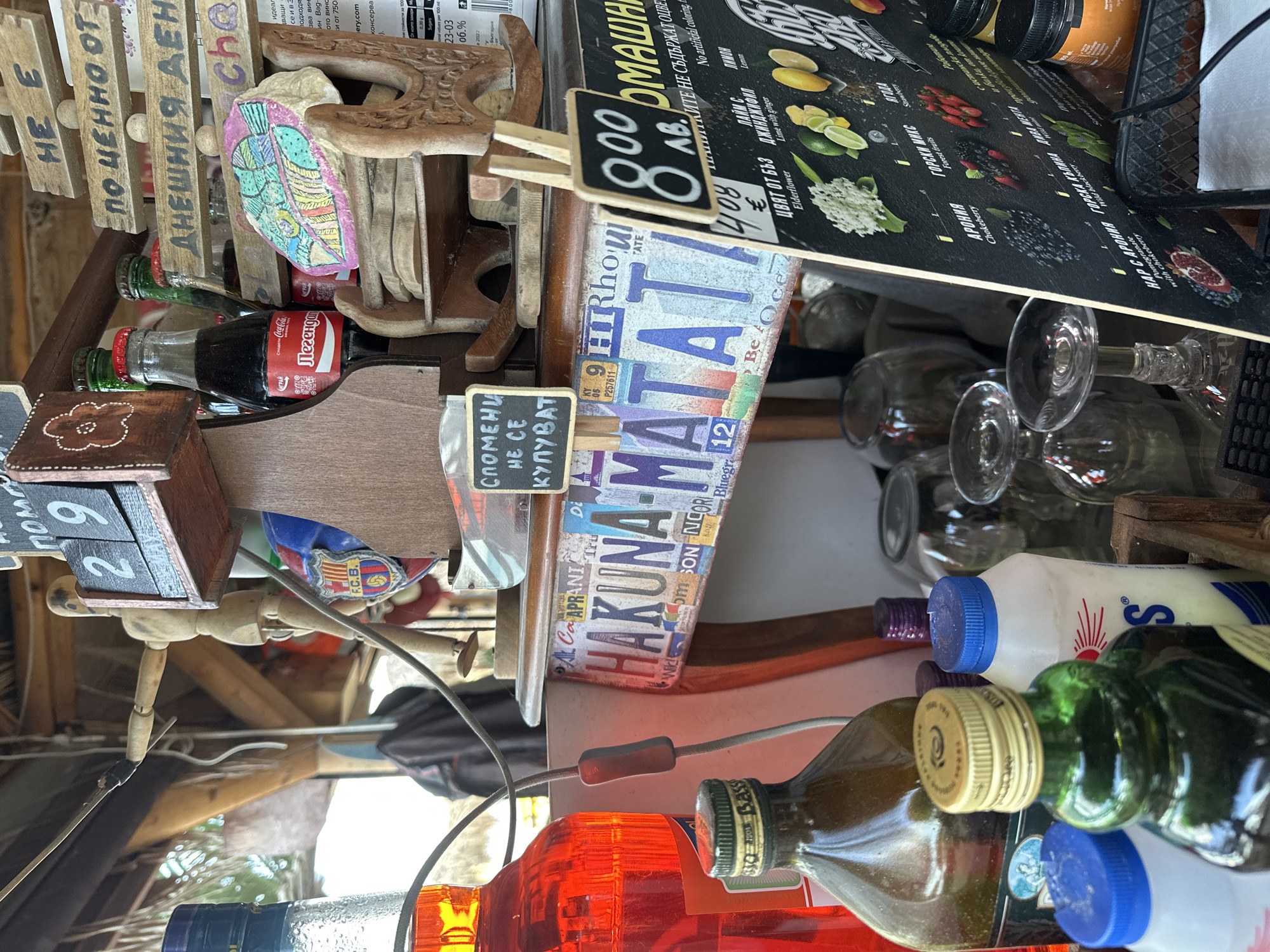 The image depicts a cluttered outdoor bar or cafe in Varna, Bulgaria. The scene is densely packed with various objects. Several glass bottles filled with colorful liquids, including red, brown, and green substances, are visible in the foreground. Above them are several clear, empty wine glasses. Adjacent to the glasses are bottles with blue, white, and yellow caps. A black sign with white text describing various beverage ingredients, such as elderflower, lime with ginger, and forest fruits, hangs above the bottles. Below the sign, a license plate with "HAKUNA MATATA" written on it is visible. Various other decorative wooden objects, including a calendar showing the number "29", small Coca-Cola bottles, and painted stones, are scattered throughout the scene. A small chalkboard with text in Bulgarian is visible. A wooden mannequin stands among the items, partially obscured by a cable.