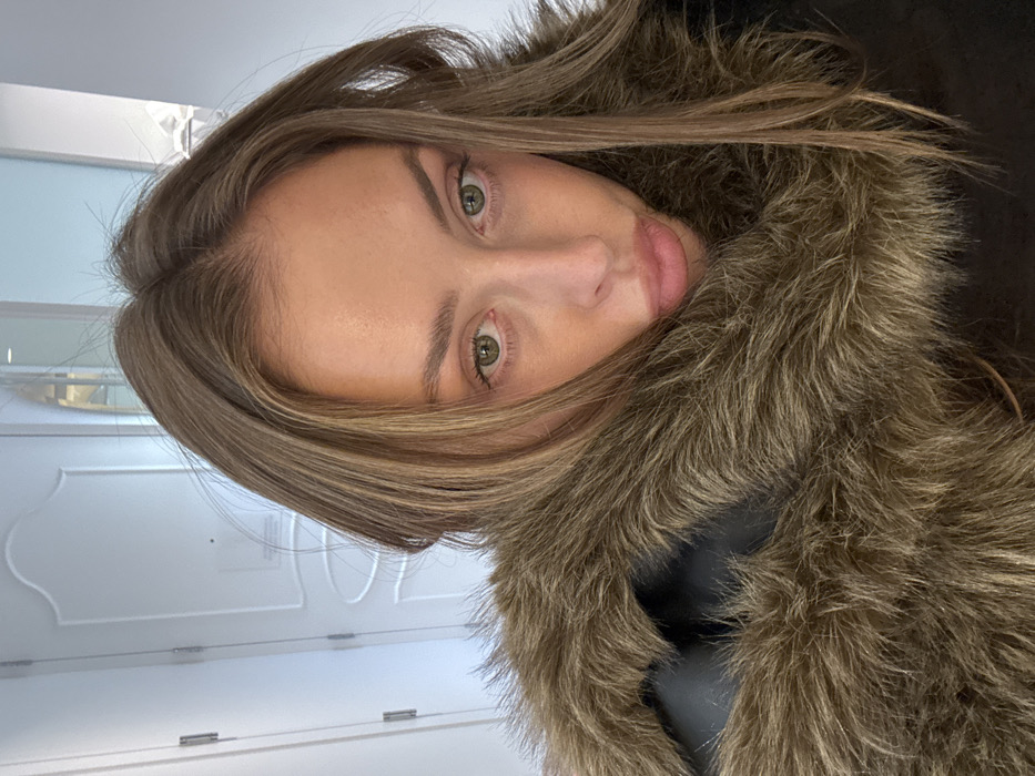 A woman with fair skin, light brown hair, and green eyes is the focus. Her hair is parted off-center. She's wearing a dark coat with a thick, fur-trimmed collar. The background features a white door with decorative molding and a hint of a mirrored surface. The overall image appears to be a selfie.
