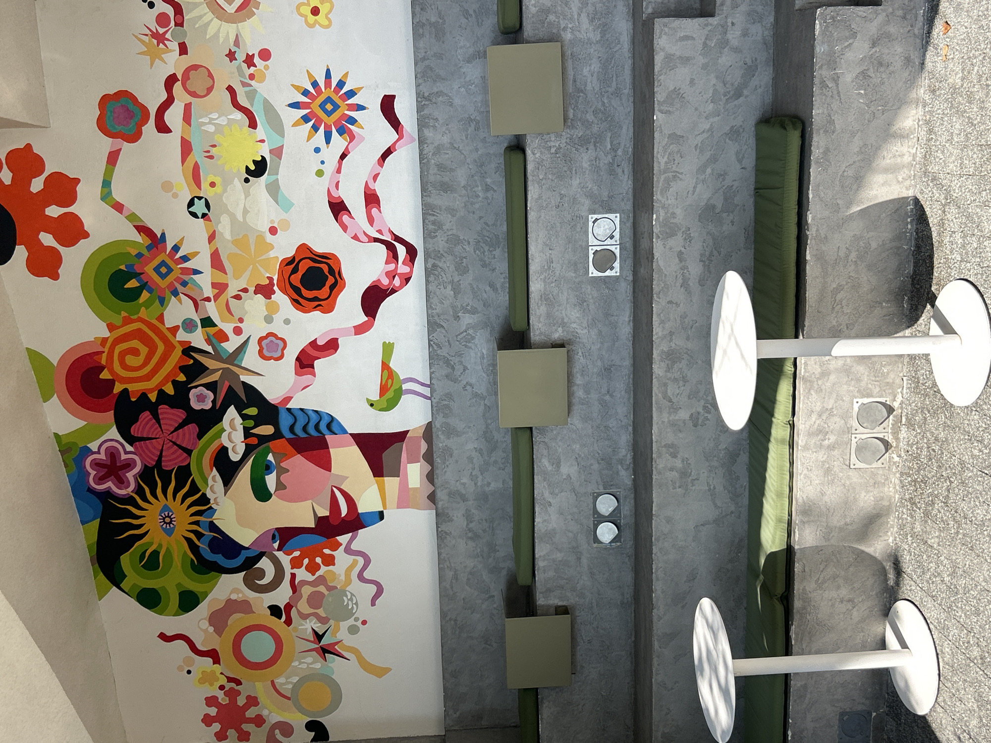 The image depicts an outdoor seating area with a colorful mural on a white wall. The mural features a stylized face with vibrant, abstract elements resembling hair, flowers, and abstract shapes. A small green and orange bird is painted to the right of the face. Below the mural, there are tiered concrete benches with green cushions. Two white circular tables with single pedestals are positioned in front of the benches. The ground is a textured gray surface. Two circular outlets are visible on the lower tier of the bench structure. The lighting suggests it is daytime, with shadows cast by the tables. This scene is located in the city of Sochi, Russia.
