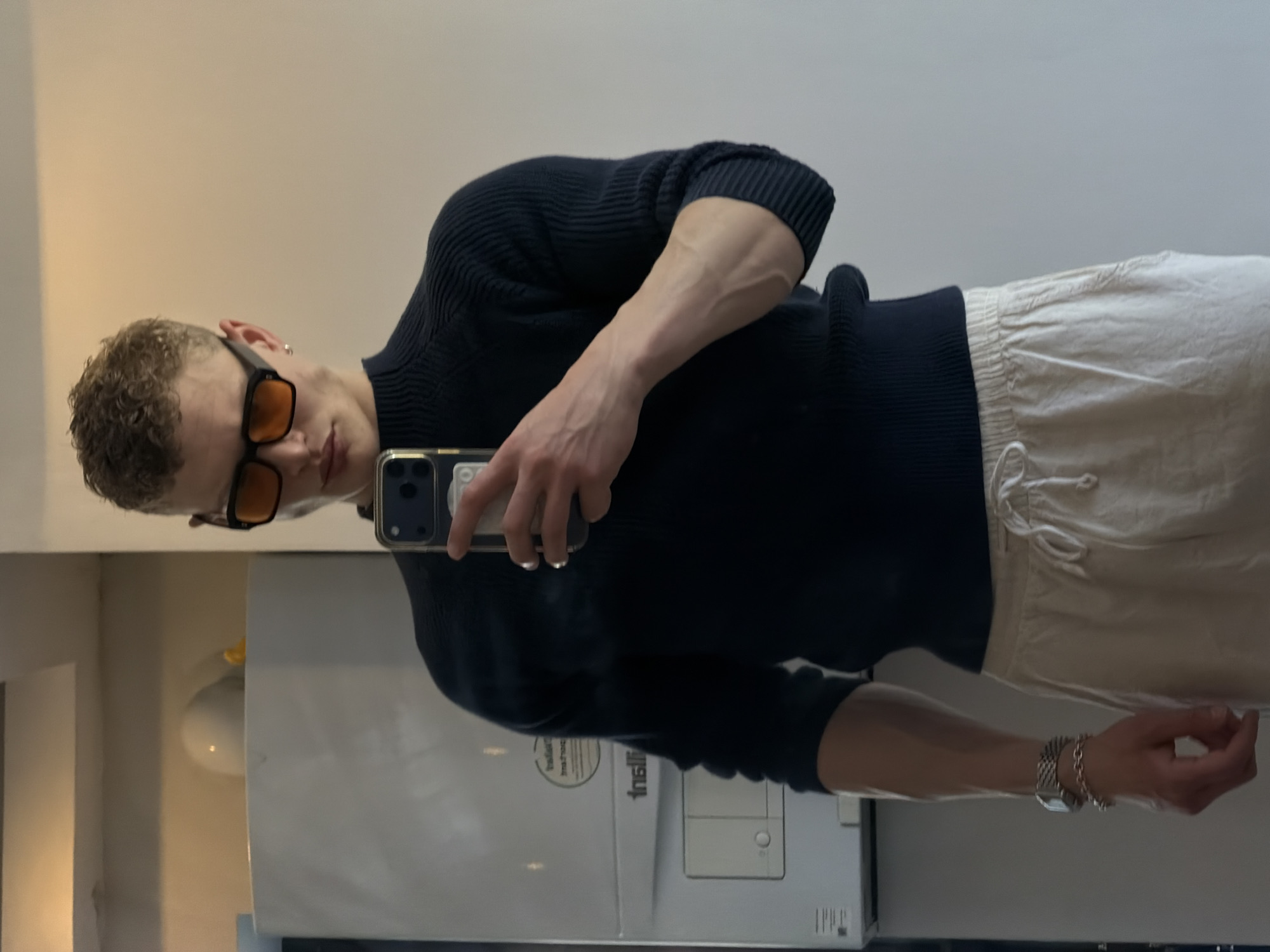 A young, light-skinned individual with short, curly blond hair is taking a selfie in front of a mirror. They are wearing orange-tinted sunglasses, a dark blue ribbed sweater, and white drawstring pants. A silver watch and multiple silver bracelets are visible on their left wrist. The individual is holding a smartphone with a clear case. The reflection in the mirror reveals a room with light-colored walls and a white appliance, possibly a refrigerator or washing machine, behind them. A small, round light fixture is visible on the wall. The location is undeterminable.