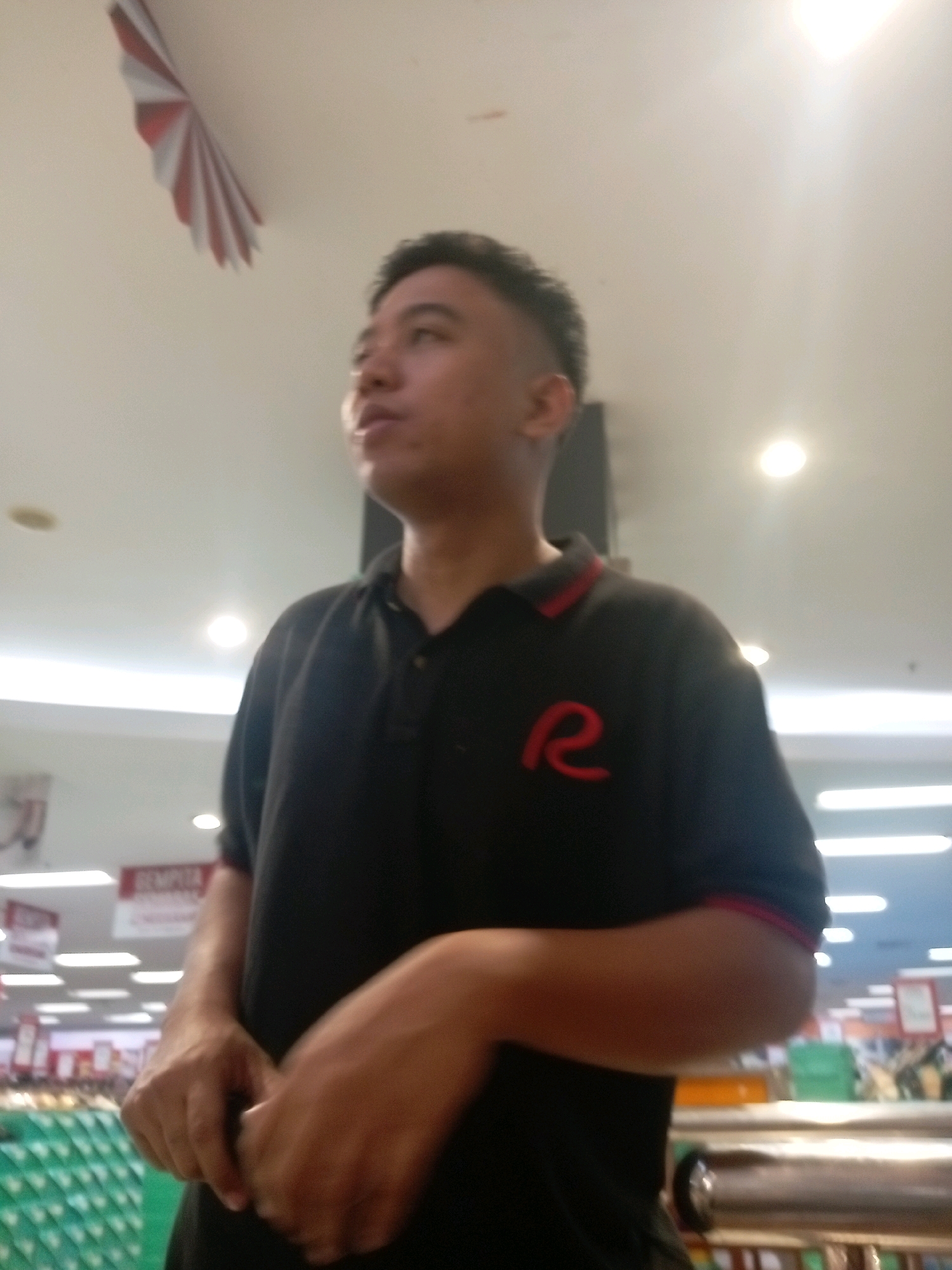 The image shows a young man wearing a black polo shirt with a red "R" logo on the chest. He is standing indoors, possibly in a retail store, with a ceiling fan and lighting visible. The man has short dark hair and is looking to his left. In the background, there are shelves stocked with items and a green basket. The overall lighting suggests an indoor environment with artificial lights. There are no clear indicators of the specific location in Batam, Indonesia, other than it being an indoor commercial space.