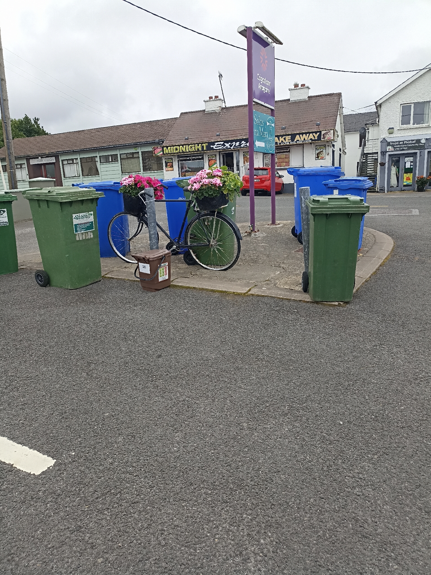 In Annagry, Ireland, a scene unfolds in front of a row of businesses including a takeaway and another establishment with a sign reading "Solas na Mara". A cluster of green and blue recycling bins is positioned on a paved area. A bicycle, adorned with baskets of pink flowers on its handlebars and rear, stands near the bins. A small brown bin, possibly for compost, is placed on the ground beside the bicycle. The sky is overcast, and the general environment appears to be a commercial or service area within the town. There are no people visible in the image.