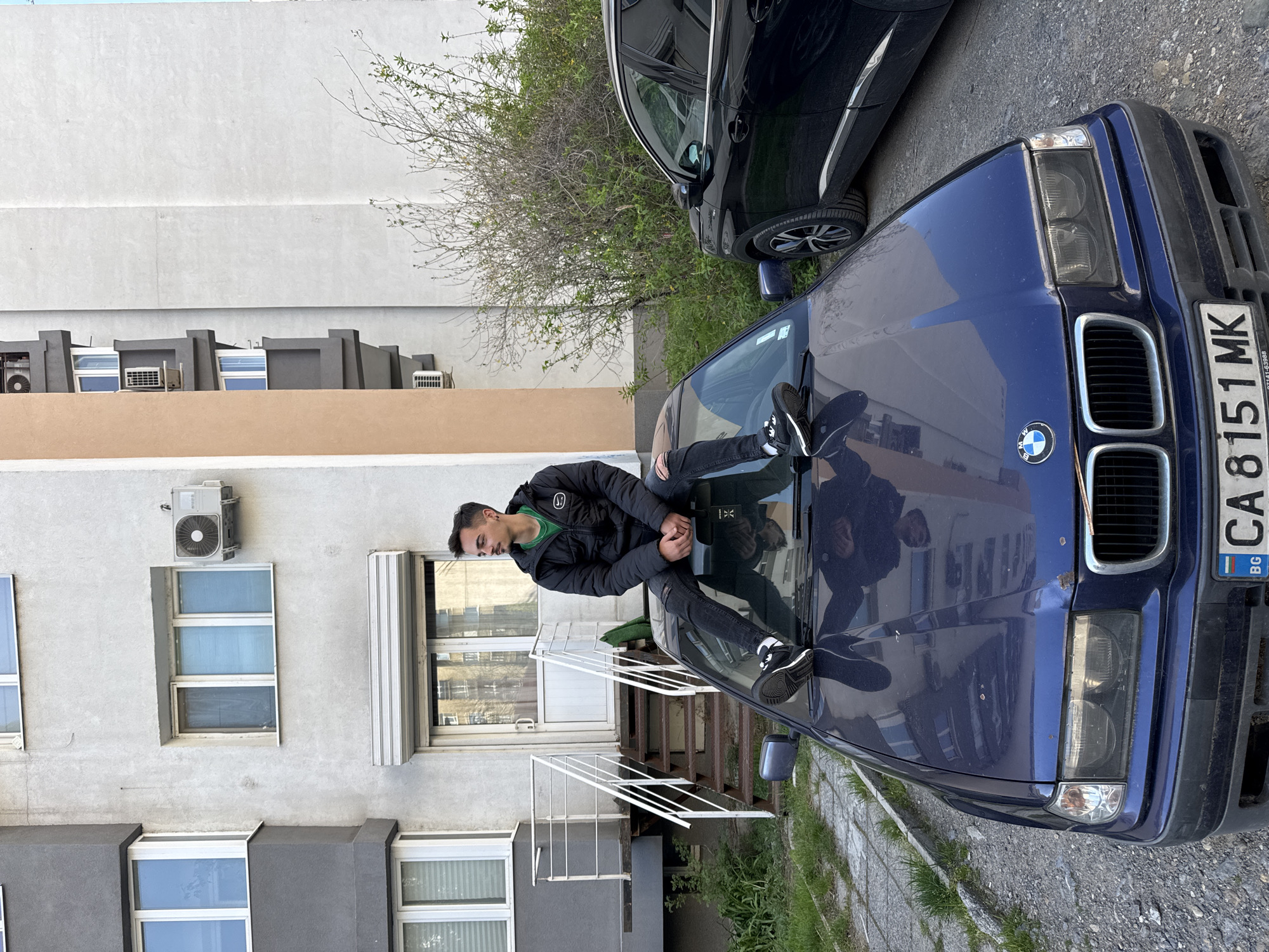 A young man is casually seated on the hood of a dark blue BMW car, facing towards the right side of the frame (if the image were rotated to be upright). He is wearing a black puffer jacket with a visible white Nike swoosh logo on the left chest, a green t-shirt, dark jeans, and black sneakers with white soles. His hands are clasped in front of him, and his reflection is clearly visible on the car's windshield.
The blue BMW, an older model (likely an E36 series), is parked on what appears to be a paved area or street. Its Bulgarian license plate "CA 8151 MK" is prominently displayed on the front bumper, along with the BMW logo on the grille. Behind the BMW, a newer, dark-colored SUV or car is partially visible.
The setting is an urban outdoor environment, with a multi-story building serving as the backdrop. The building features light gray or off-white walls, with some sections of darker gray and a reddish-brown strip. Several windows and external air conditioning units are affixed to the building's facade. Bare tree branches with some emerging green leaves are visible near the top right of the frame. The ground beneath the cars consists of a mix of pavement, gravel, and sparse grass.
The scene is illuminated by bright daylight, suggesting a clear day, possibly late morning or afternoon, with distinct shadows cast on the car and ground. The overall atmosphere appears casual and relaxed. The location is confirmed as Sofia, Bulgaria, by the license plate.