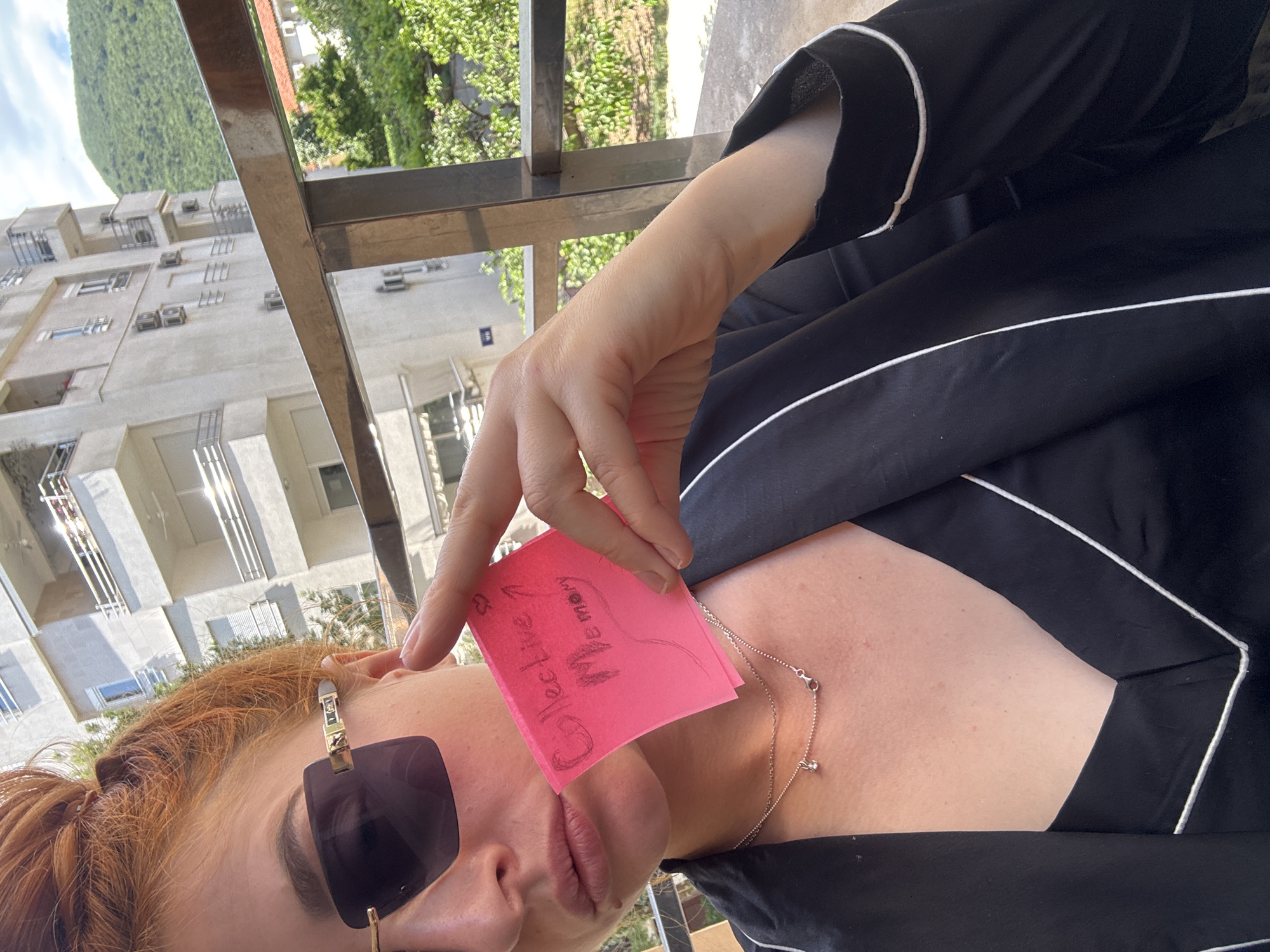 A woman stands on a balcony in Budva, Montenegro, holding a pink sticky note with the handwritten text "Collective Memory" over her face. She is wearing dark sunglasses and a black top with white piping. A silver necklace is visible around her neck. The balcony has a metal railing. In the background is a multi-story residential building and a green hillside or mountain. The weather appears sunny.