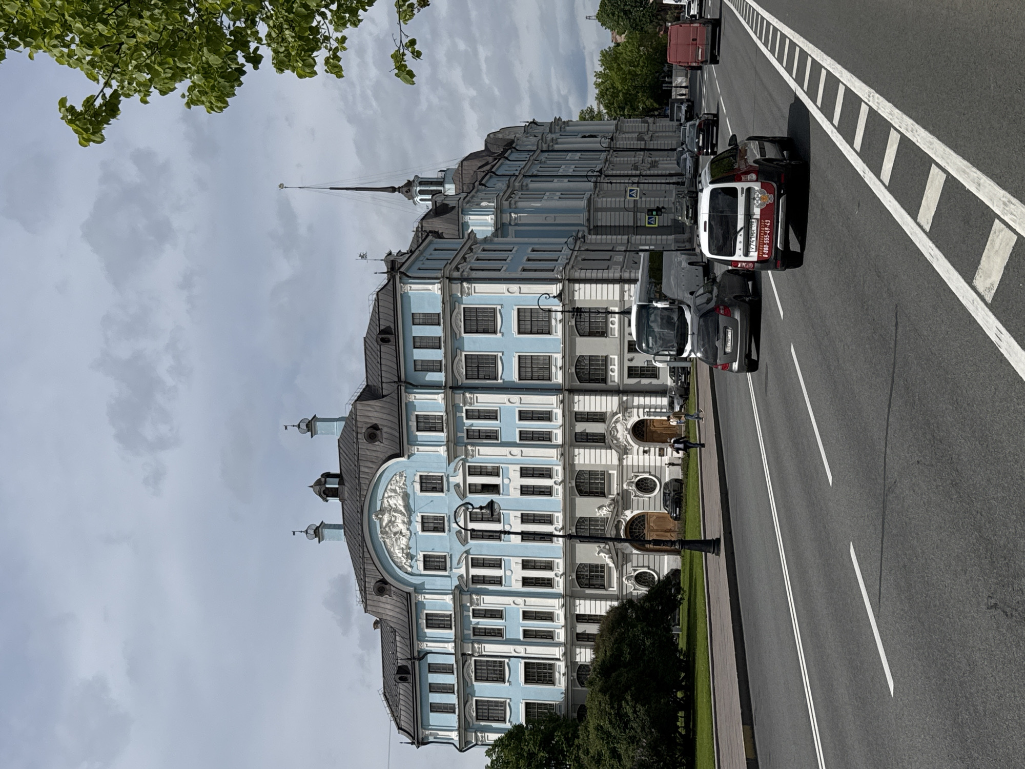 A street scene in Saint Petersburg, Russia, captured on an overcast day, features a grand, historic building alongside a multi-lane asphalt road. The prominent building, appearing to be the former Senate and Synod or a similar Neoclassical/Baroque structure, boasts a light blue and white facade, dark grey mansard roofs, and several ornate spires, including a slender, tall spire topped with a golden orb visible against the cloudy sky. Its architecture is rich with details such as white pilasters, arched windows, decorative elements, and a sculptural frieze above what may be a main entrance.
To the right of the building, a wide road with white solid, dashed, and block-like markings indicates multiple lanes and a pedestrian crossing. Several vehicles are visible, including cars and vans. A white delivery van prominently displays Cyrillic text: "ЛАДА ЭКСПРЕСС" (Lada Express) and "МОЙКА ОКОН" (Window Washing) along with a phone number "8-800-555-49-40" and a Russian license plate "Y774 MP 98," with the "98" region code confirming Saint Petersburg. Other vehicles, like a red truck and various dark cars, are also present.
Green foliage from trees frames the upper left corner of the image, and a line of trees runs along a paved sidewalk that separates the building from the road. A few indistinct human figures are visible near the building's entrances and walking on the sidewalk. The overall impression is that of a bustling city street with impressive historical architecture.