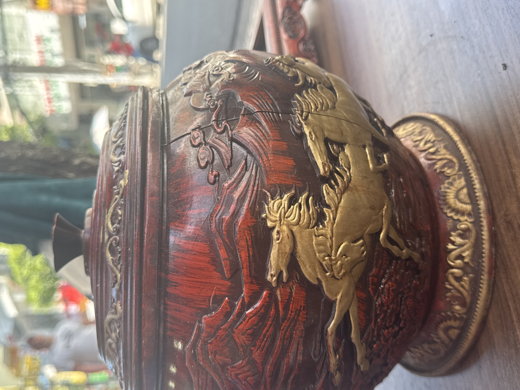 A close-up shot shows a dark red and gold decorative vessel with a horse motif. The vessel is resting on a wooden surface. The background is blurred, showing a cityscape through a window. The location is Unknown, Unknown.