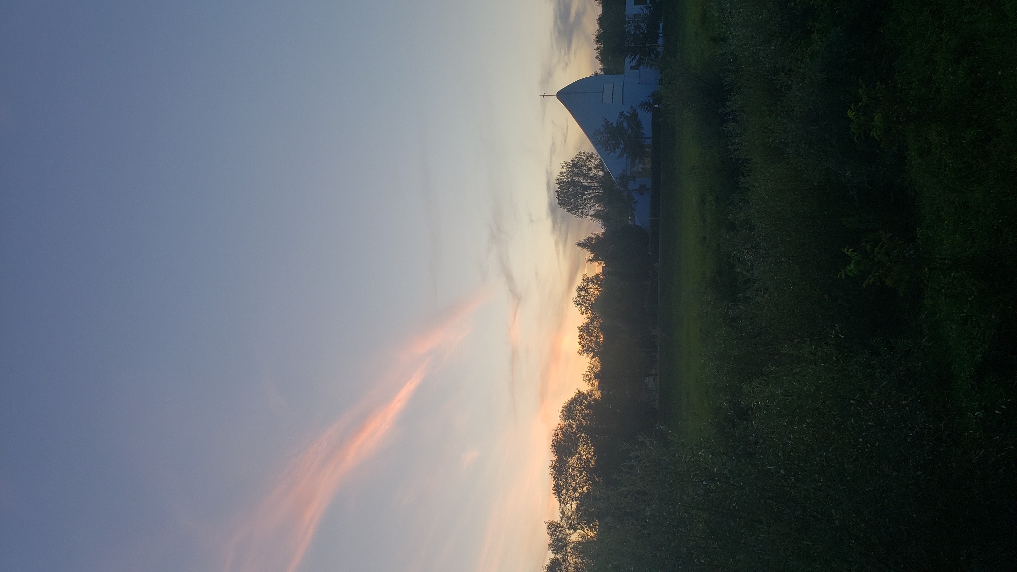 The image depicts a view of the Church of the Holy Spirit in Prešov, Slovakia, during what appears to be sunset or early dawn. The sky is a gradient of soft blues and grays, with streaks of pale pink clouds stretching across the upper left portion of the frame. In the middle ground, a line of dark green trees forms a silhouette against the lighter sky. To the right of the trees, the distinct architectural shape of the Church of the Holy Spirit is visible. It is a modern structure with a prominent, pointed roof, painted in a light blue hue, and topped with a cross. The church is surrounded by more trees and what appears to be some buildings. The foreground is dominated by a dense, dark green area of vegetation, likely a field or park, with some smaller bushes and trees visible. The overall scene is serene and quiet, capturing the twilight atmosphere of the city.