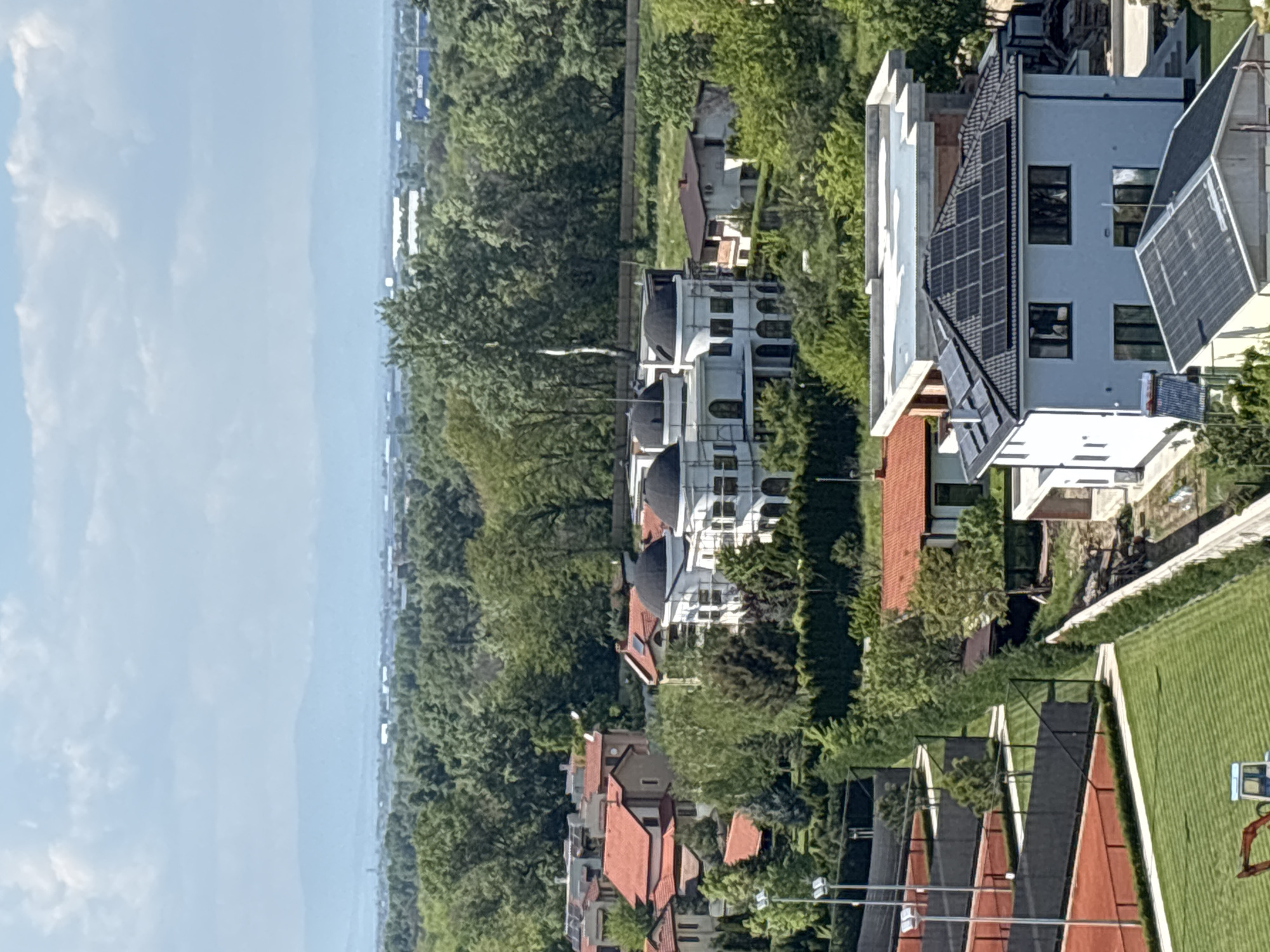 The image shows a high-angle view of a residential area, possibly in Plovdiv, Bulgaria. The foreground features a row of houses with red-tiled roofs and well-manicured lawns. Some houses have solar panels installed on their roofs. A building with white walls and domed roofs is also visible. The background consists of dense trees and a distant cityscape under a light blue sky with clouds.