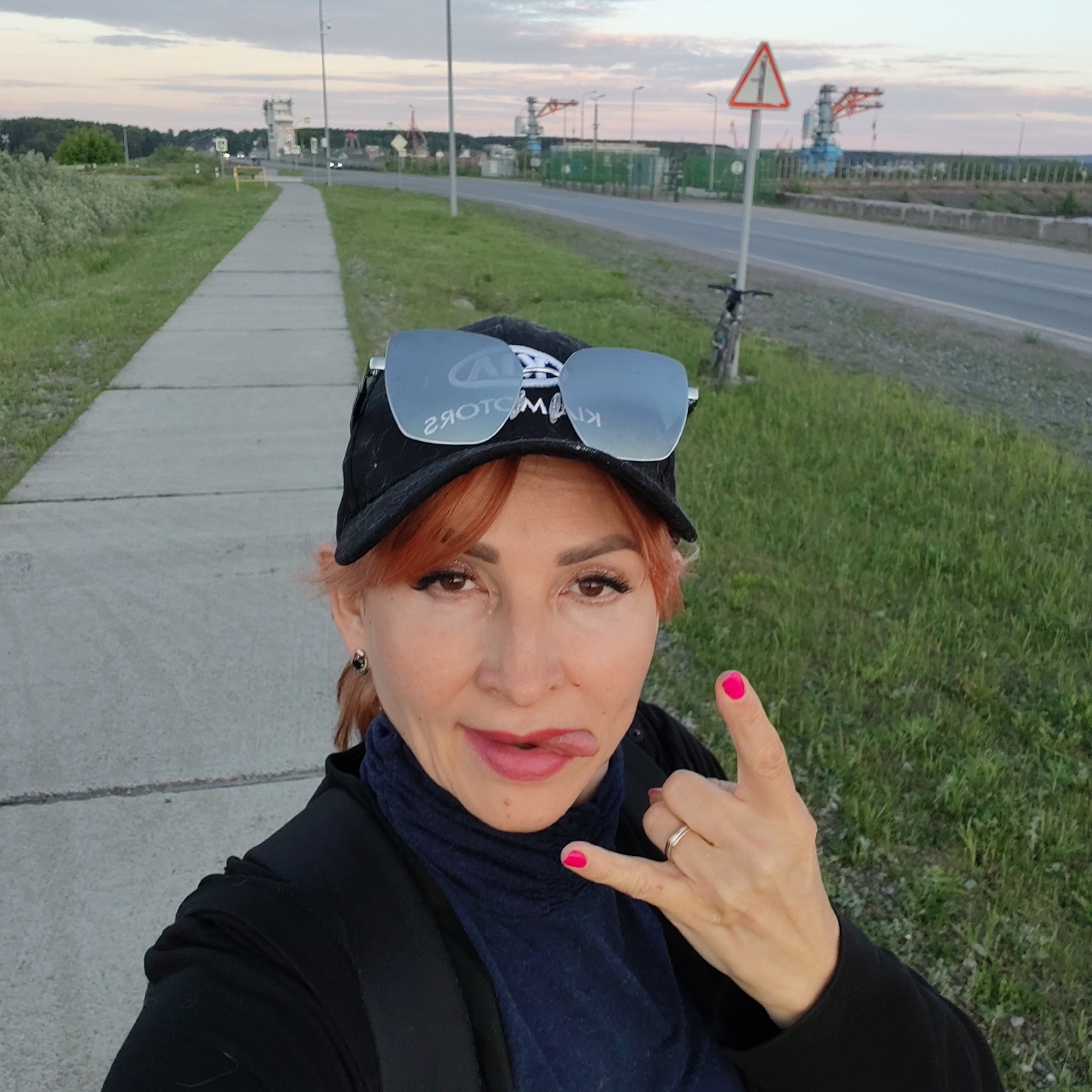 A woman takes a selfie outdoors in what appears to be the evening or late afternoon, given the soft, slightly pink hues in the sky. She has reddish-brown hair, dark eyes, and a playful expression with her tongue slightly out. She is wearing a black baseball cap with large, mirrored sunglasses pushed up on top. The reflection in the sunglasses clearly shows the reversed text "KIA MOTORS." Her right hand is making a "rock and roll" gesture (corna), and her fingernails are painted a bright pink. She wears a silver ring on her right ring finger. Her attire includes a dark blue turtleneck or mock neck top and a black jacket.
The scene behind her features a concrete sidewalk to her left and a paved road to her right, separated by green grass. In the distance, along the road, there are industrial structures, possibly cranes or parts of a port facility, consistent with the location context of Chaikovsky, Russia, which is on the Kama River. A white triangular road sign with a red border and a black exclamation mark inside (indicating "Danger" or "Other dangers") is visible on the right side of the road, close to a bicycle leaning against a pole. Utility poles and streetlights line the road, and some small green portable structures or containers are also seen. The sky is largely overcast but shows some light at the horizon. The overall impression is a casual moment captured in an industrial or semi-industrial outdoor setting.