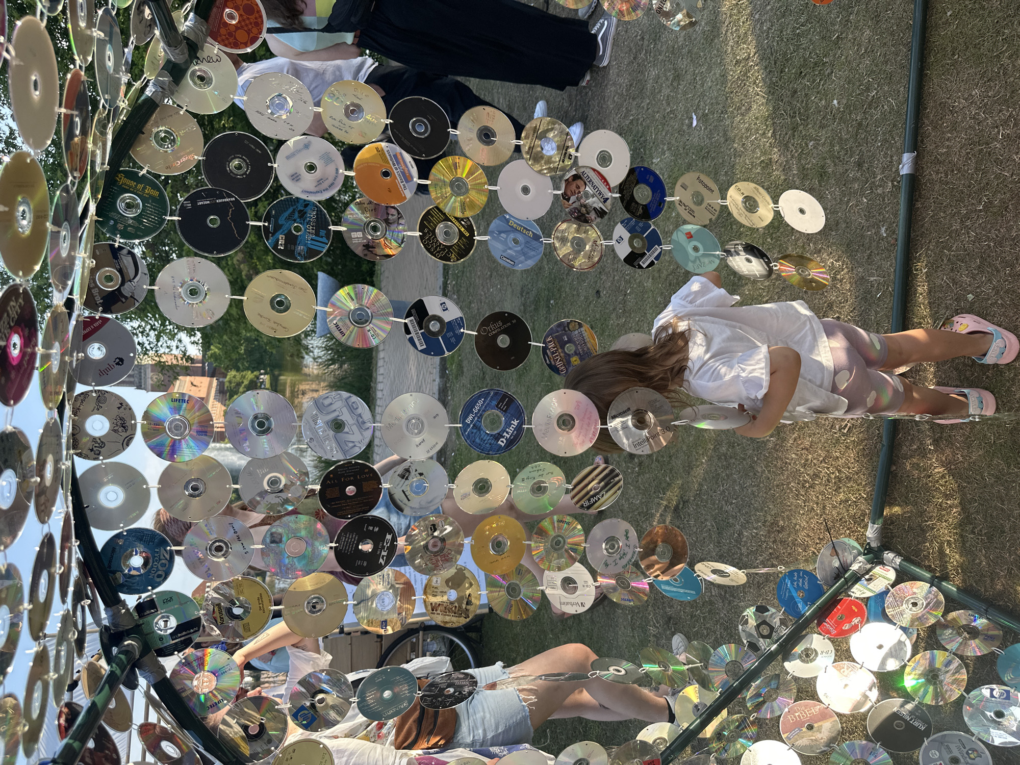 The image depicts an outdoor art installation made of numerous CDs, strung together and suspended from a green metal frame. The CDs are arranged in vertical columns and rows, creating a shimmering, reflective curtain. The scene is set in a grassy area, with a body of water and buildings visible in the background, suggesting a park or public space.
In the foreground, a young child with long brown hair, wearing a white t-shirt and patterned shorts, stands facing away from the camera, looking at the CD installation. Their feet are clad in colorful slide sandals. To the left of the frame, partially visible, is a person wearing denim shorts and a white top, with a brown fanny pack around their waist. Further in the background, other individuals are present, blurred and out of focus, some wearing dark clothing.
The CDs themselves vary in color and design, with some displaying printed labels or artwork, and others reflecting the sunlight and surroundings. The overall impression is a whimsical and creative use of recycled materials, likely at an event or festival.
The prompt specifies the location as the city of Kiel, Germany. Considering the context of outdoor art installations and public spaces in Kiel, this scene is likely located at the **former Kieler Seegarten (Kiel Sea Garden)**, a popular venue that has hosted various art exhibitions and cultural events, particularly during the summer months. The presence of water and open green space aligns with the characteristics of this location.