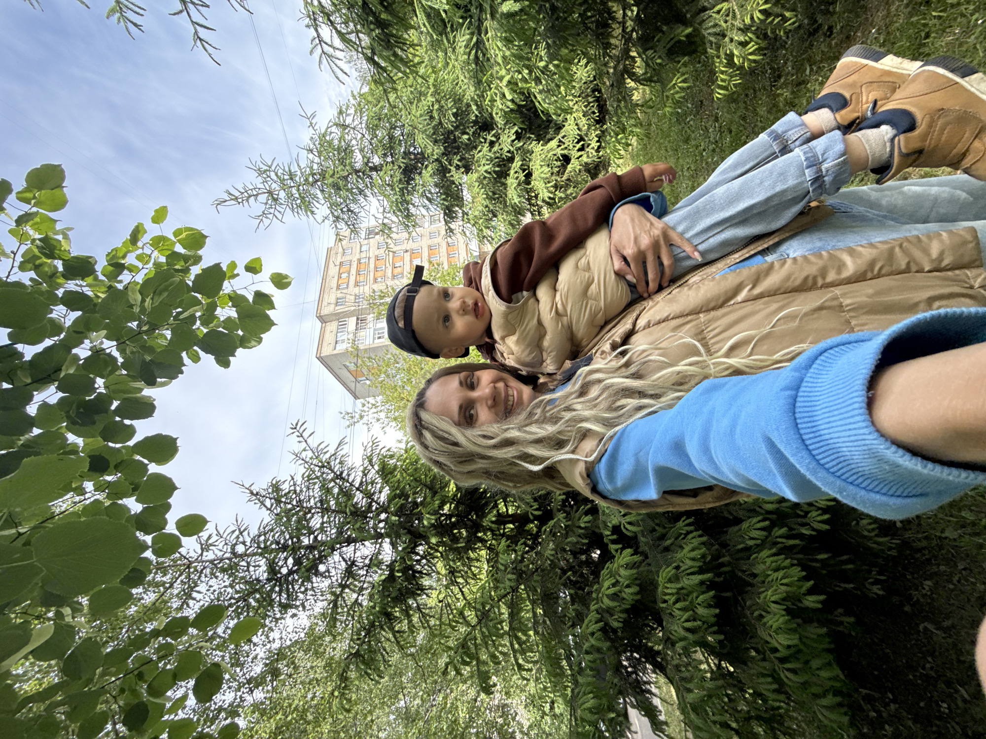 The photograph depicts a woman and a toddler outdoors, possibly in a park setting. The woman, wearing a blue long-sleeved shirt and a brown puffer vest, is holding the toddler on her lap. The toddler is wearing a brown shirt, a tan vest, and a black baseball cap. They are positioned against a backdrop of trees and sky. In the background, a building with numerous windows is visible. The image is taken from a low angle, suggesting the photographer is lying down.
While Rusanovo, Russia, is mentioned, specific location confirmation within the city isn't discernible from the image. The scene features a mother and child in a public outdoor space with trees, buildings, and sky visible.